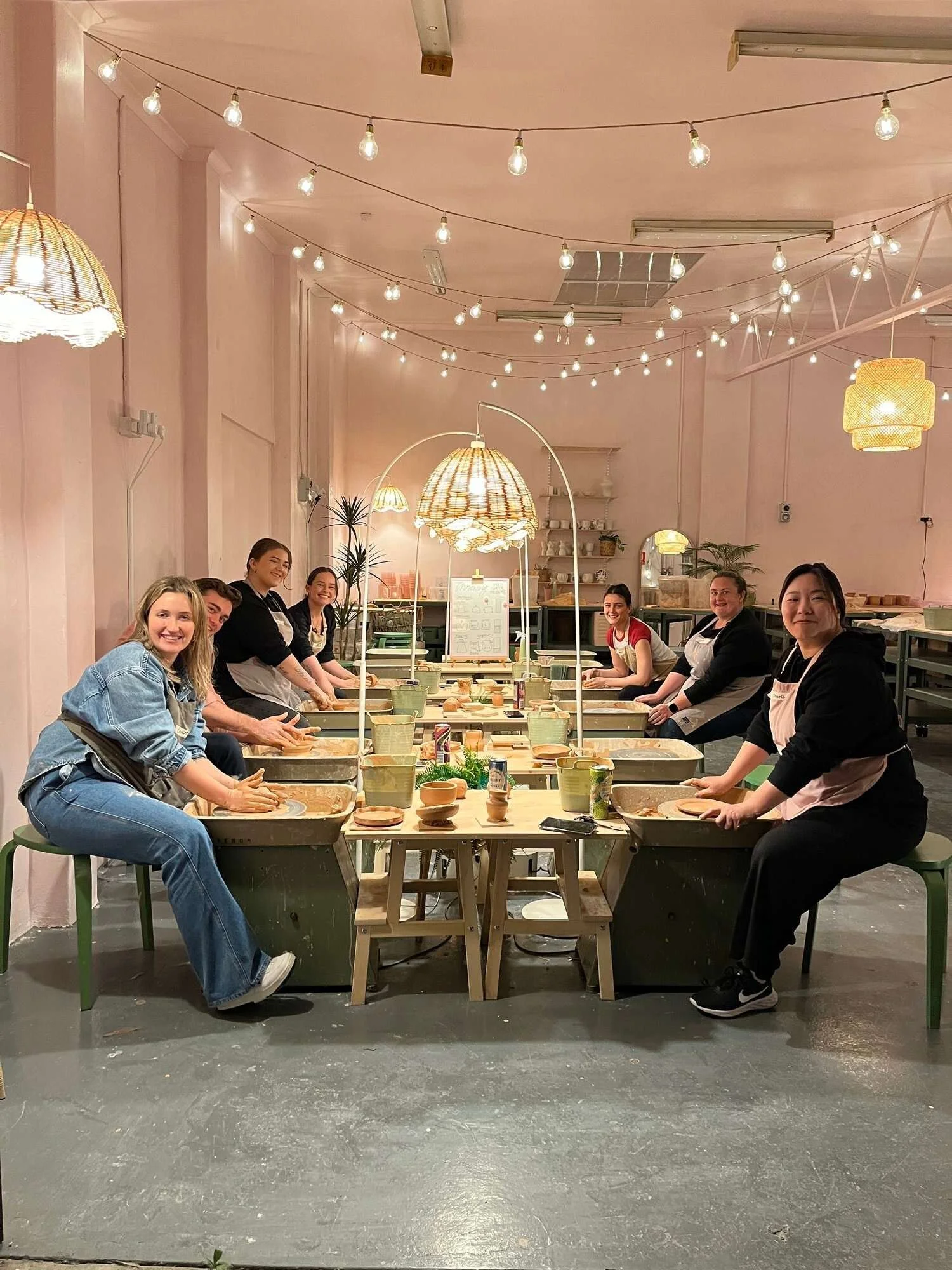 Group of eight people sitting around a pottery wheel table in a softly lit, pink-colored room with hanging lights and lamps, engaged in ceramics or pottery making activity.