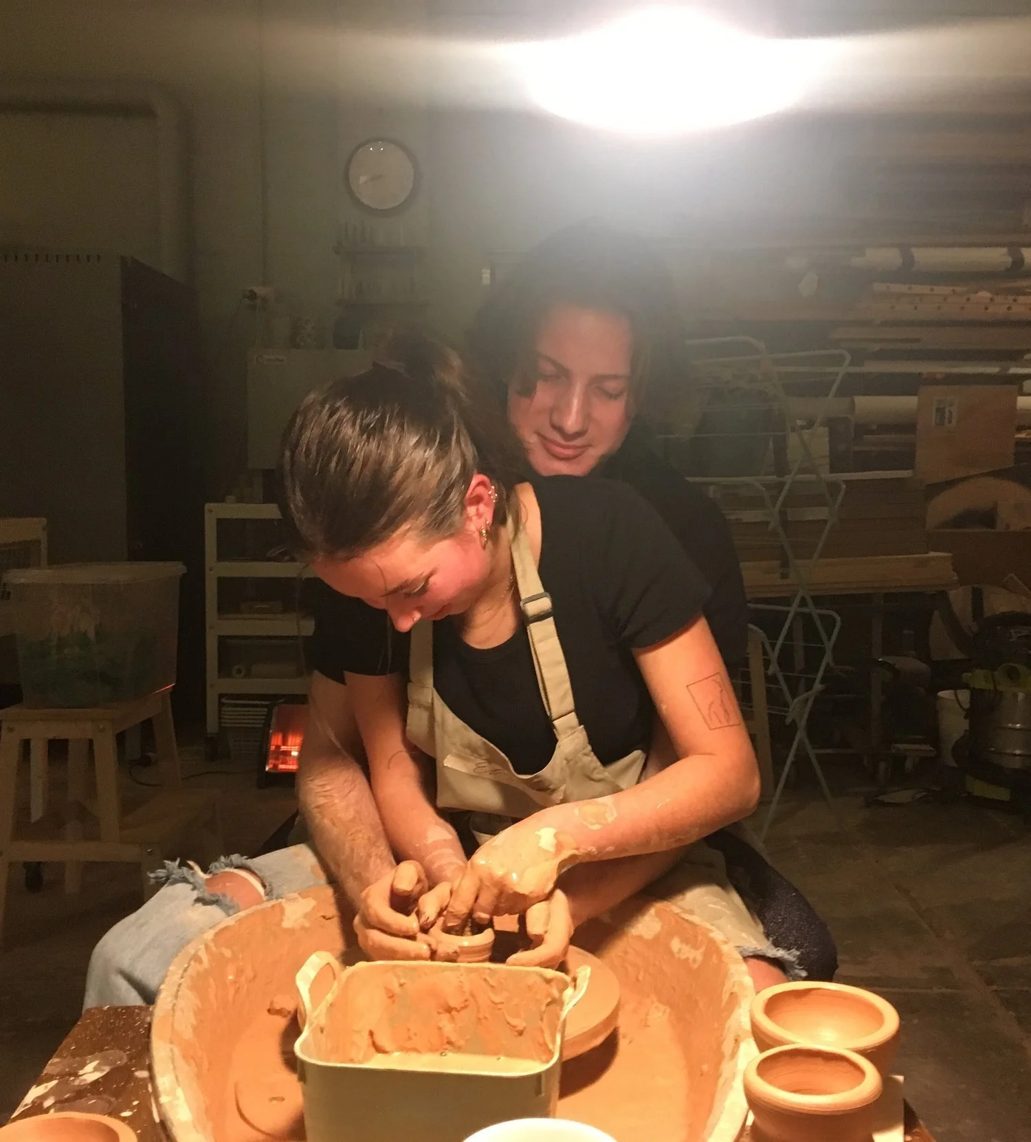 Two women are working together on a pottery wheel in a studio, shaping clay. One woman, wearing a beige apron, is focused on the pottery, while the other woman stands behind her, guiding and assisting.