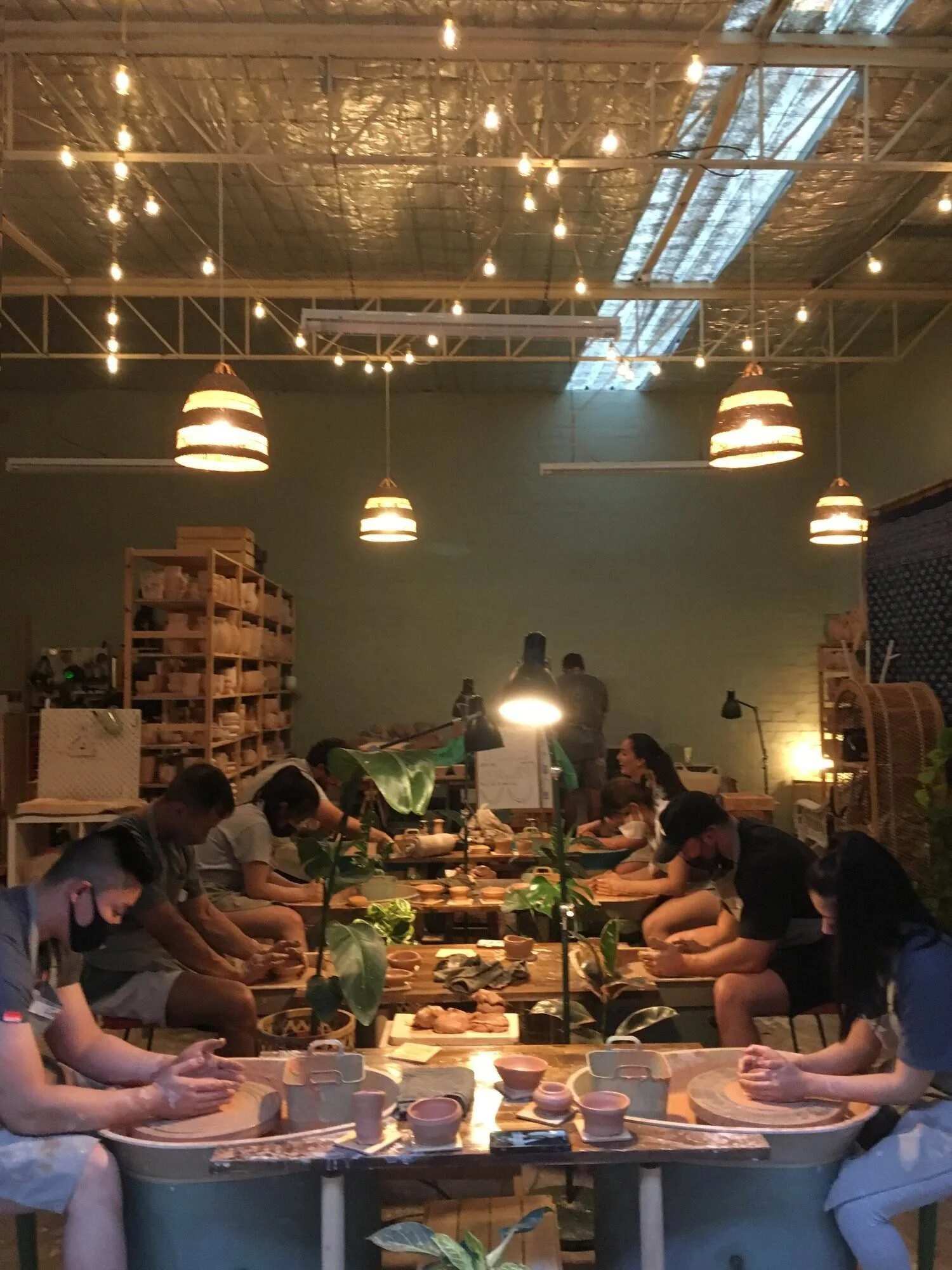 People sit at pottery wheels in a pottery studio, working on clay projects with instructors guiding them. The space is decorated with plants and shelves filled with supplies, illuminated by warm lighting and hanging pendant lights.