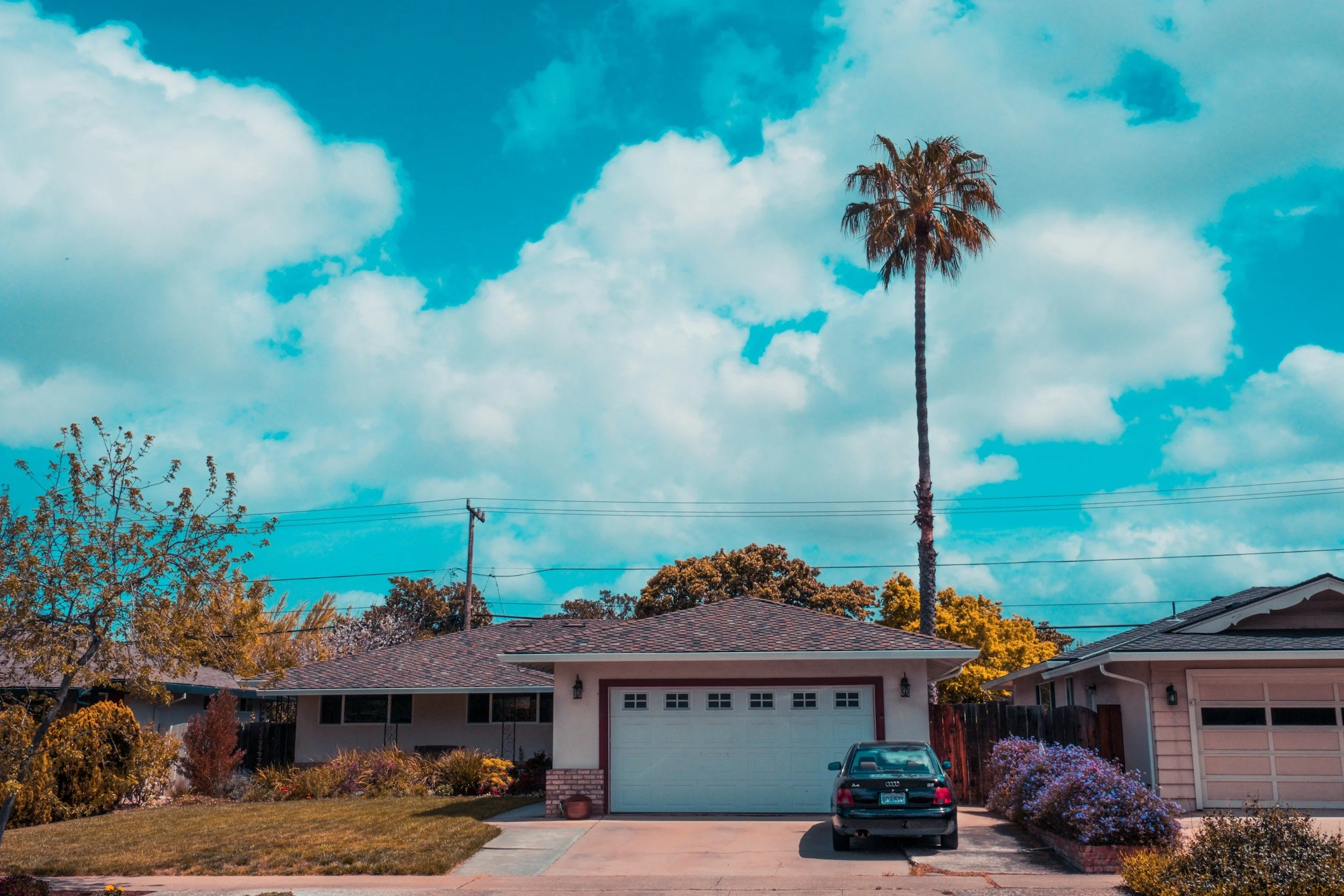 A suburban house with a garage, a car parked in the driveway, a tall palm tree, and a partly cloudy sky