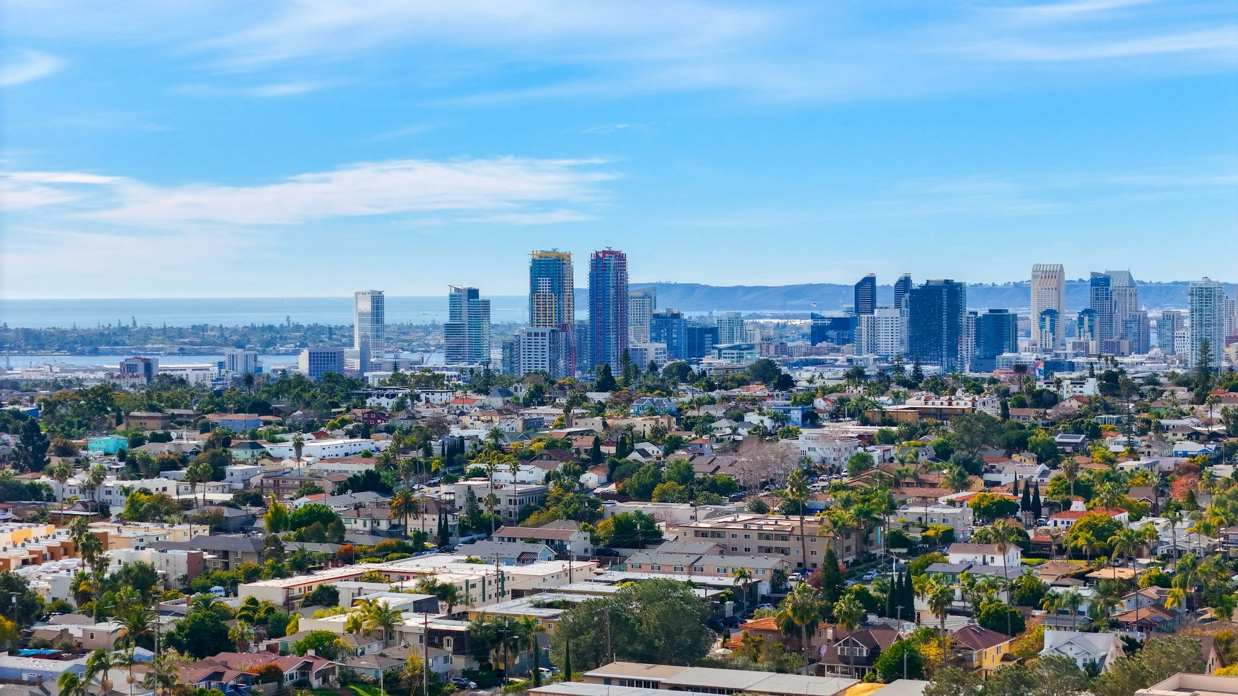 A city skyline with tall modern buildings in the background, and a residential area with houses and trees in the foreground under a blue sky with some clouds.