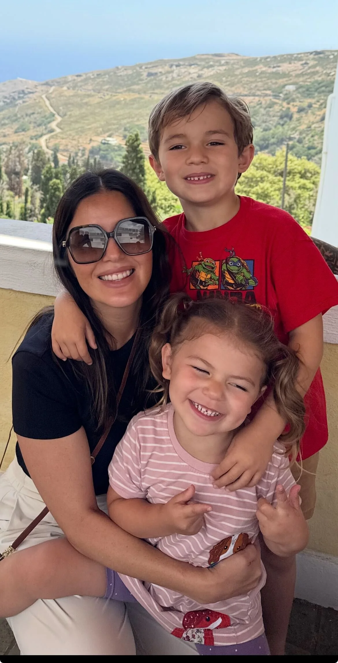 A woman with long dark hair, wearing large sunglasses and a black shirt, is smiling and surrounded by two children. A boy in a red Teenage Mutant Ninja Turtles shirt is standing behind her, and a young girl in a pink striped shirt with a strawberry pattern is in her arms, both kids are smiling and pointing. They are on a balcony with scenic hills, trees, and a distant view of the coast in the background.
