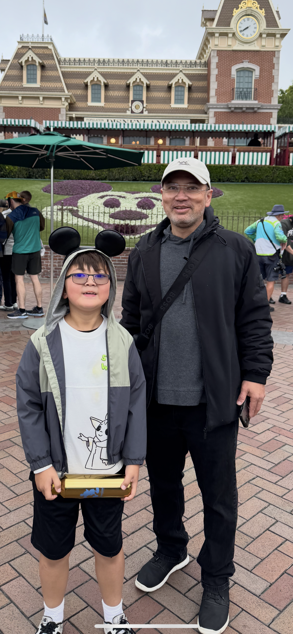 A young boy wearing Mickey Mouse ears and a gray jacket stands next to an adult man in black clothing at Disneyland. The Disneyland train station is visible in the background with a floral Mickey Mouse face on the lawn.