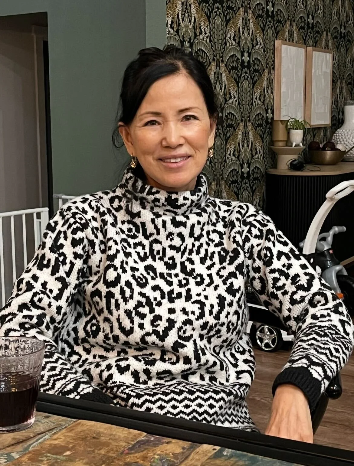 A woman with short black hair sitting at a table with a drink, wearing a black and white animal print turtleneck sweater, smiling at the camera in a cozy decorated room.