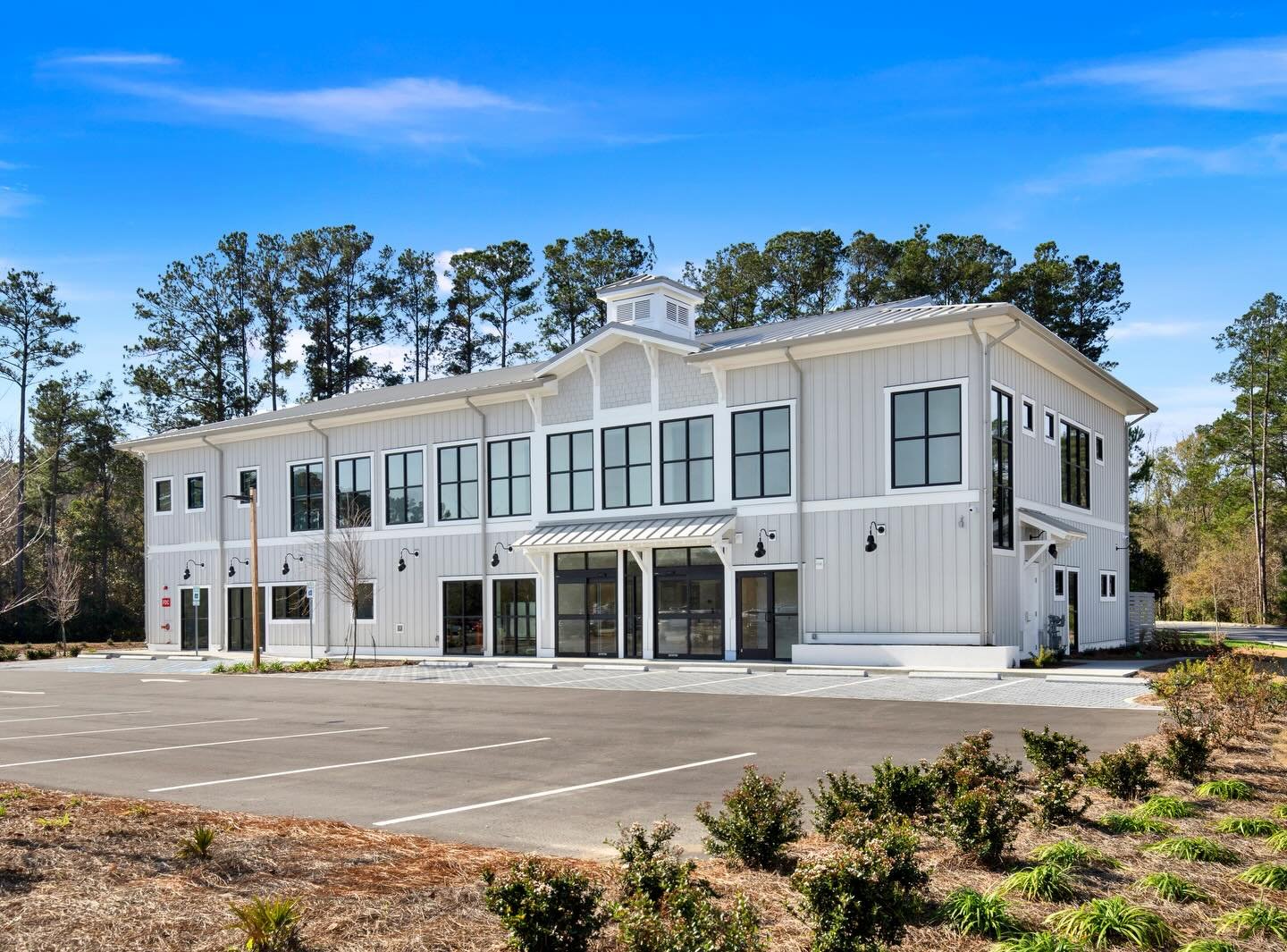 A modern white two-story building with black-framed windows and a parking lot in front, surrounded by trees under a blue sky.