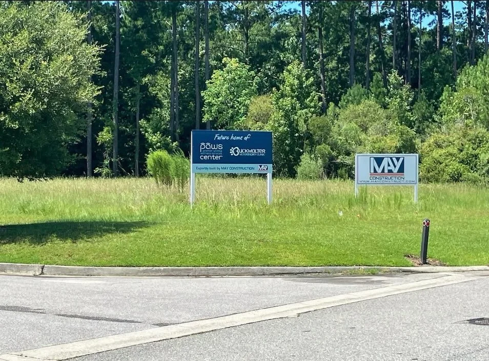 Empty lot with two construction signs and a small green bush. Sign on the left indicates future home of Paw's Center and Urgent Care Clinic by May Construction. Sign on the right displays May Construction logo.