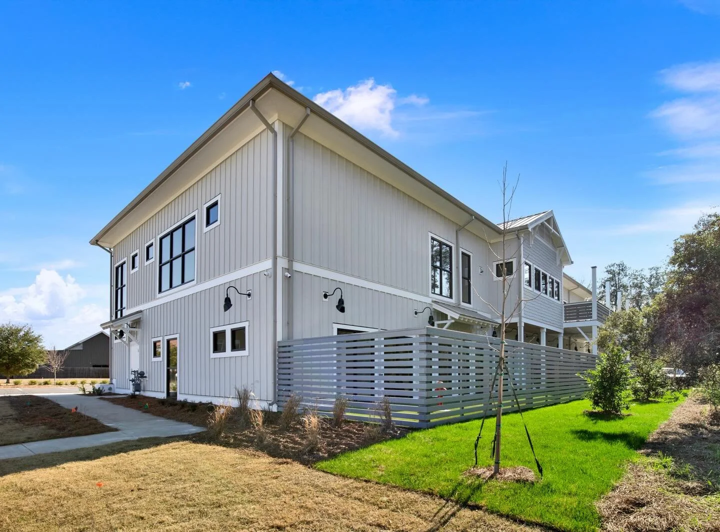 A modern two-story white house with black window frames, a landscaped yard, a young tree, a fenced patio, and a clear blue sky.