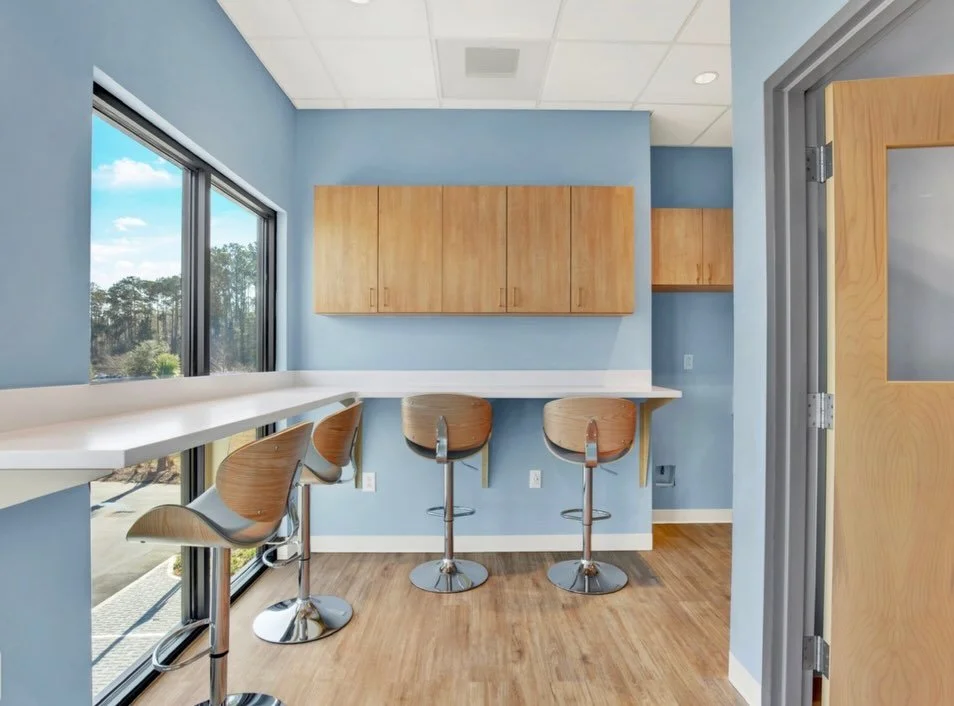 A small seating area with a blue wall, large window, wooden cabinets, a white counter, and three wooden bar stools with chrome bases.