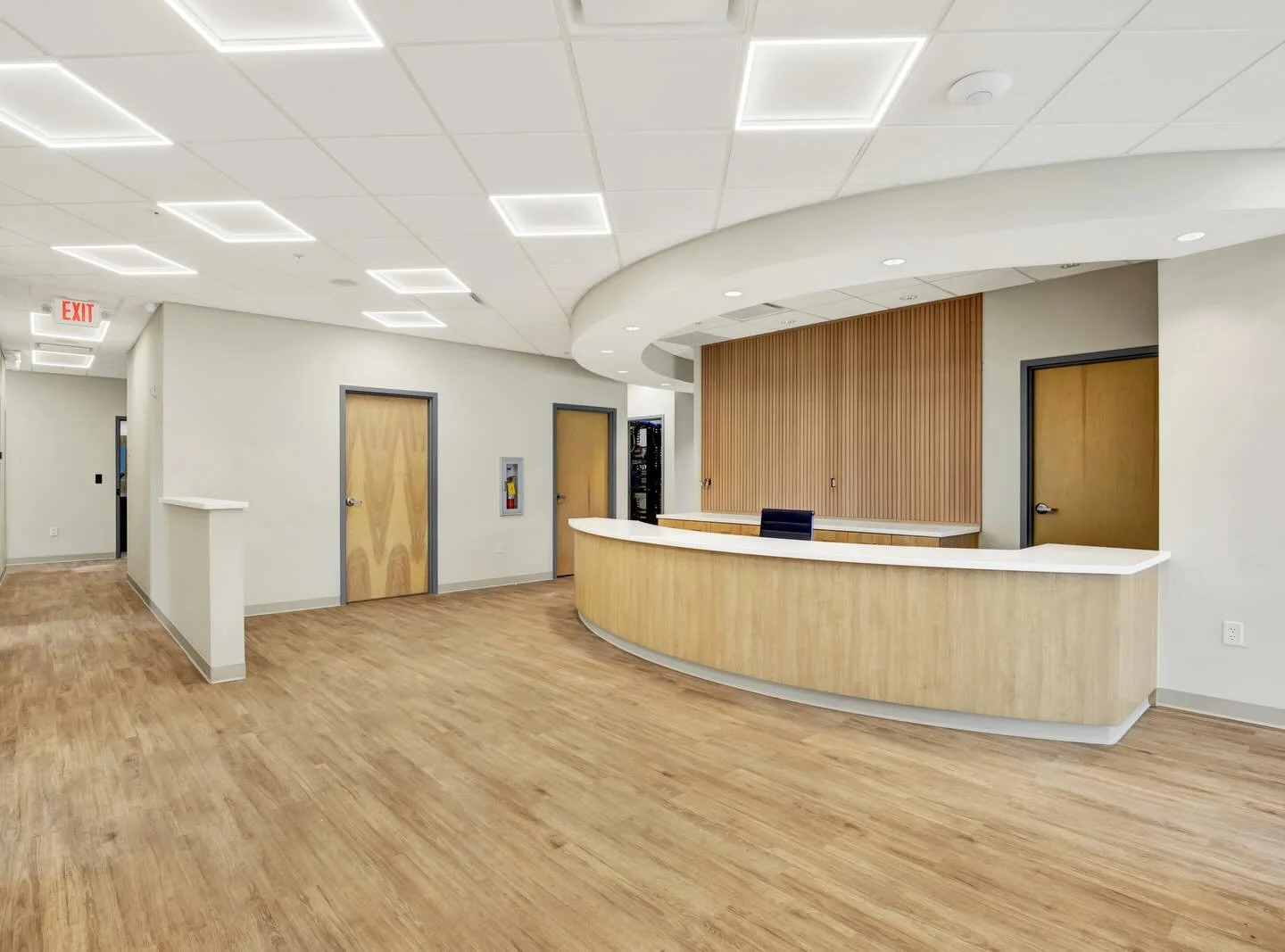 Empty reception area with a curved wooden desk, wooden flooring, and light-colored walls. There are two doors behind the desk, and ceiling lights embedded in a grid pattern. An exit sign is visible in the background.