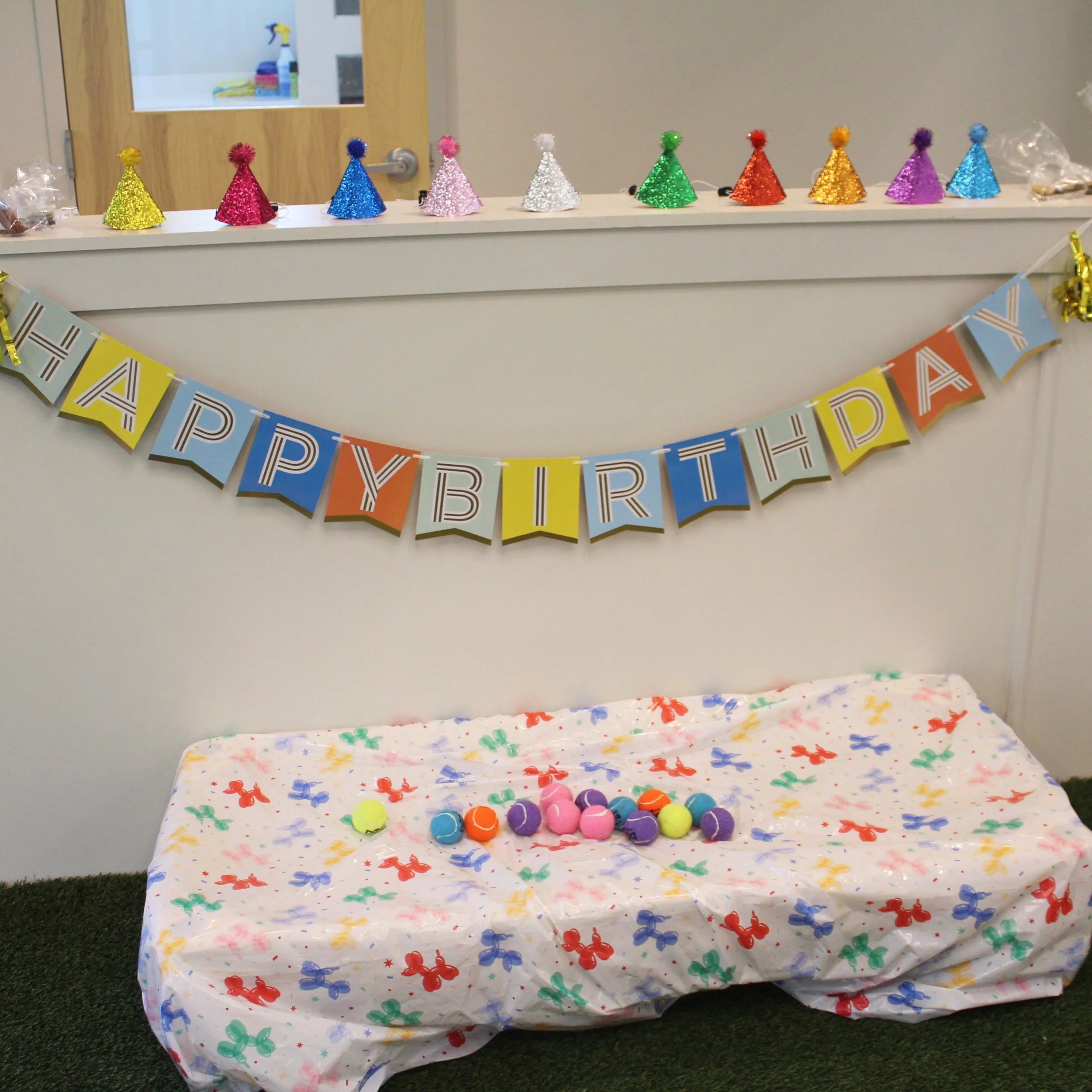 Colorful party hats with pom-poms are lined up on a table behind a festive banner that says 'Happy Birthday'. The table in front is covered with a white tablecloth decorated with multicolored balloon and star patterns, holding a collection of small, 