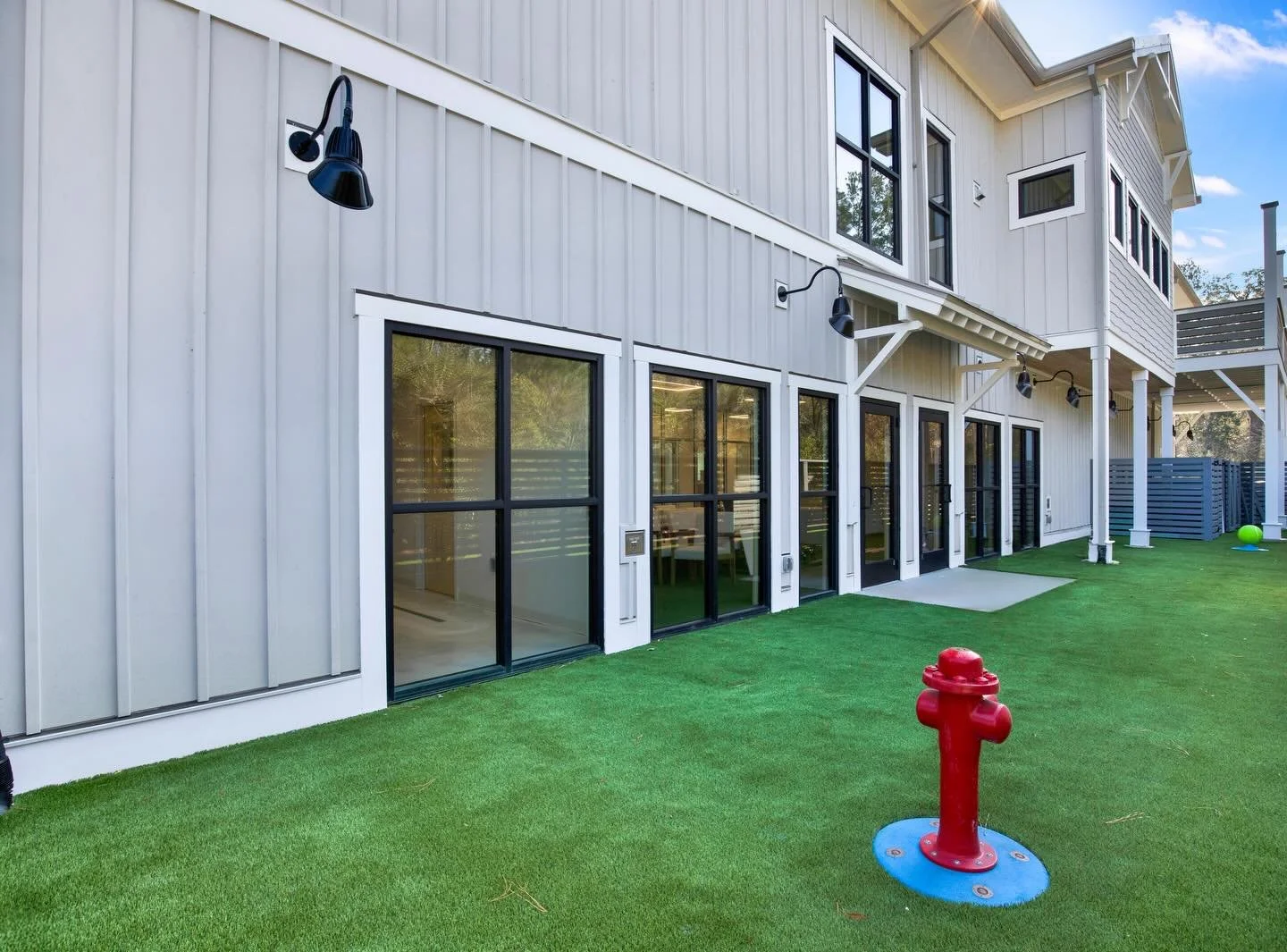 Rear view of a modern white house with large sliding glass doors, black window frames, and outdoor lighting fixtures. The house has a small balcony, and the backyard features artificial green grass with a red fire hydrant, a green ball, and a blue ba
