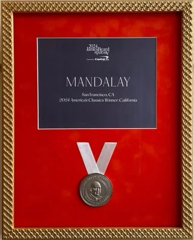 A framed award plaque with a red background, a silver medal with a 'V' above it, and a printed card indicating the 2024 James Beard Award for Best New Restaurant for Mandalay in San Francisco, California.