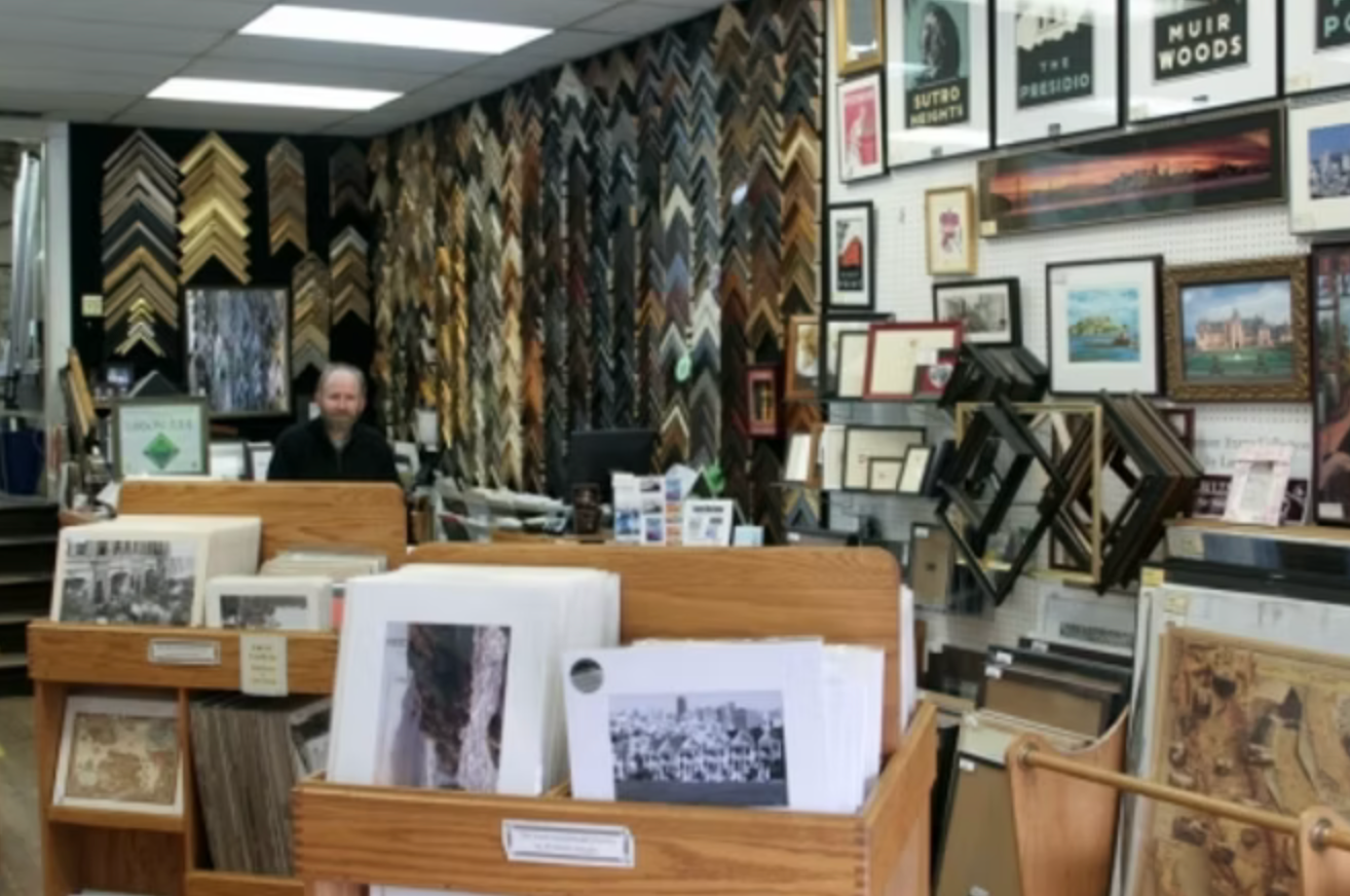 A store interior with framed artwork and picture frames on display, with a man sitting behind a counter.