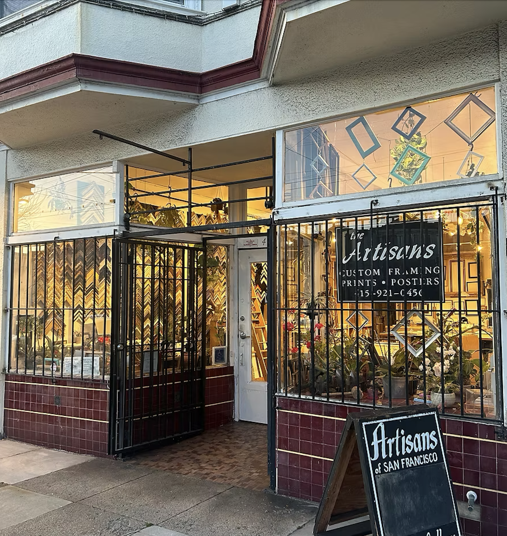 Exterior view of Artisans of San Francisco storefront with large window display, potted plants, and a black signboard outside. The shop offers custom framing, prints, and posters.