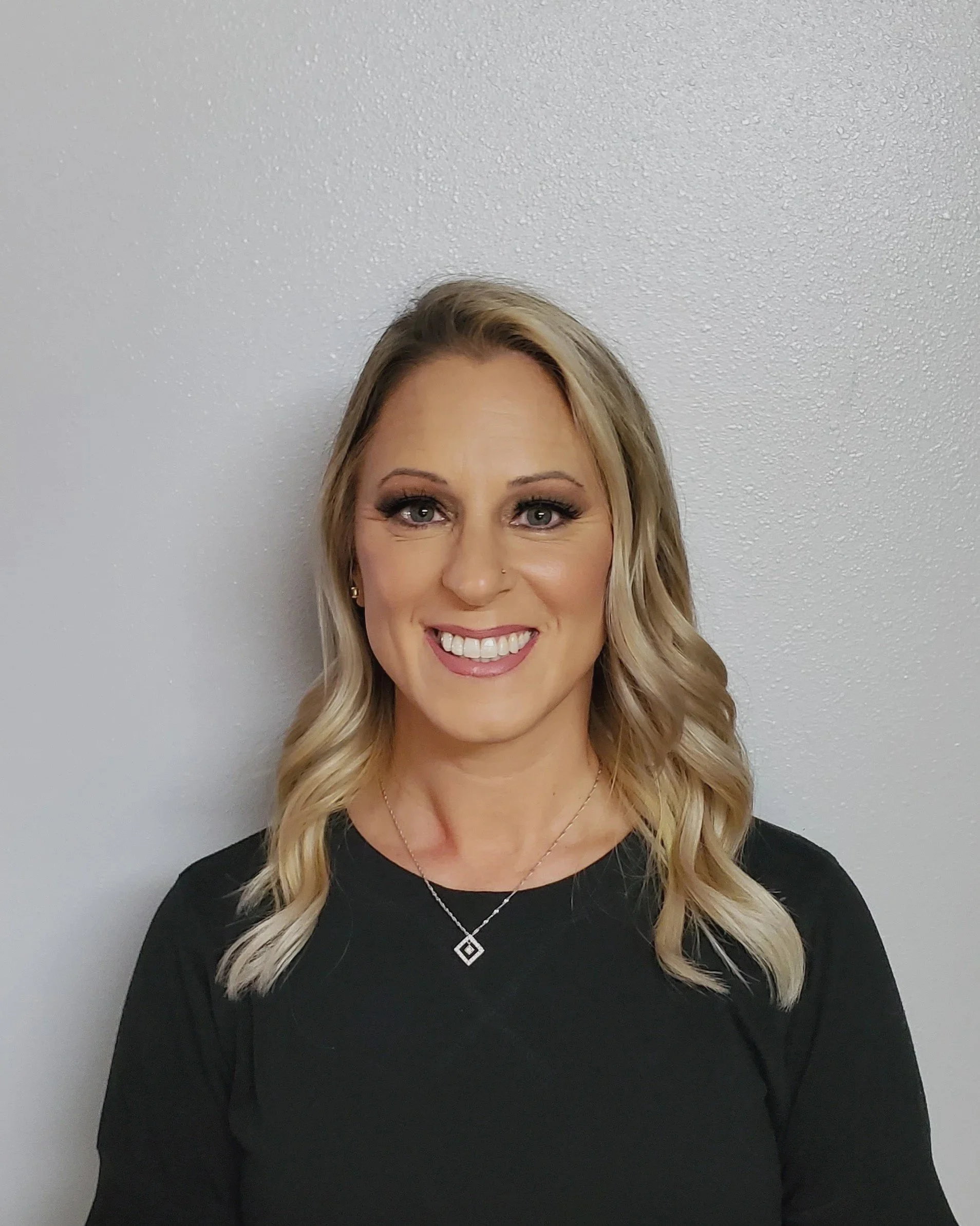 Portrait of a smiling woman with blonde wavy hair, wearing a black top and jewelry, standing against a plain, light-colored wall.