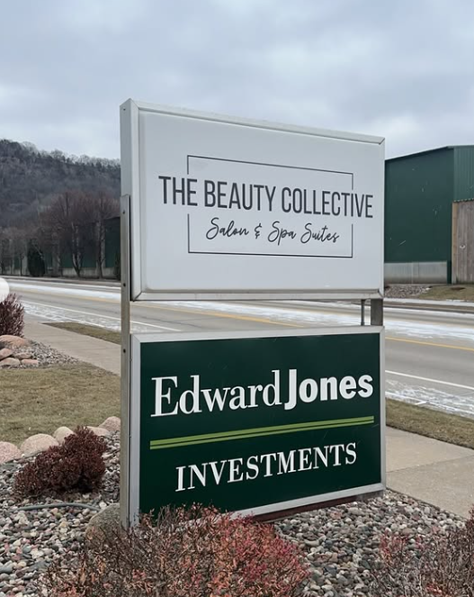 Outdoor business sign with two panels advertising The Beauty Collective salon and spa suites and Edward Jones Investments, located along a road with a sidewalk, some rocks and bushes, and a cloudy sky.
