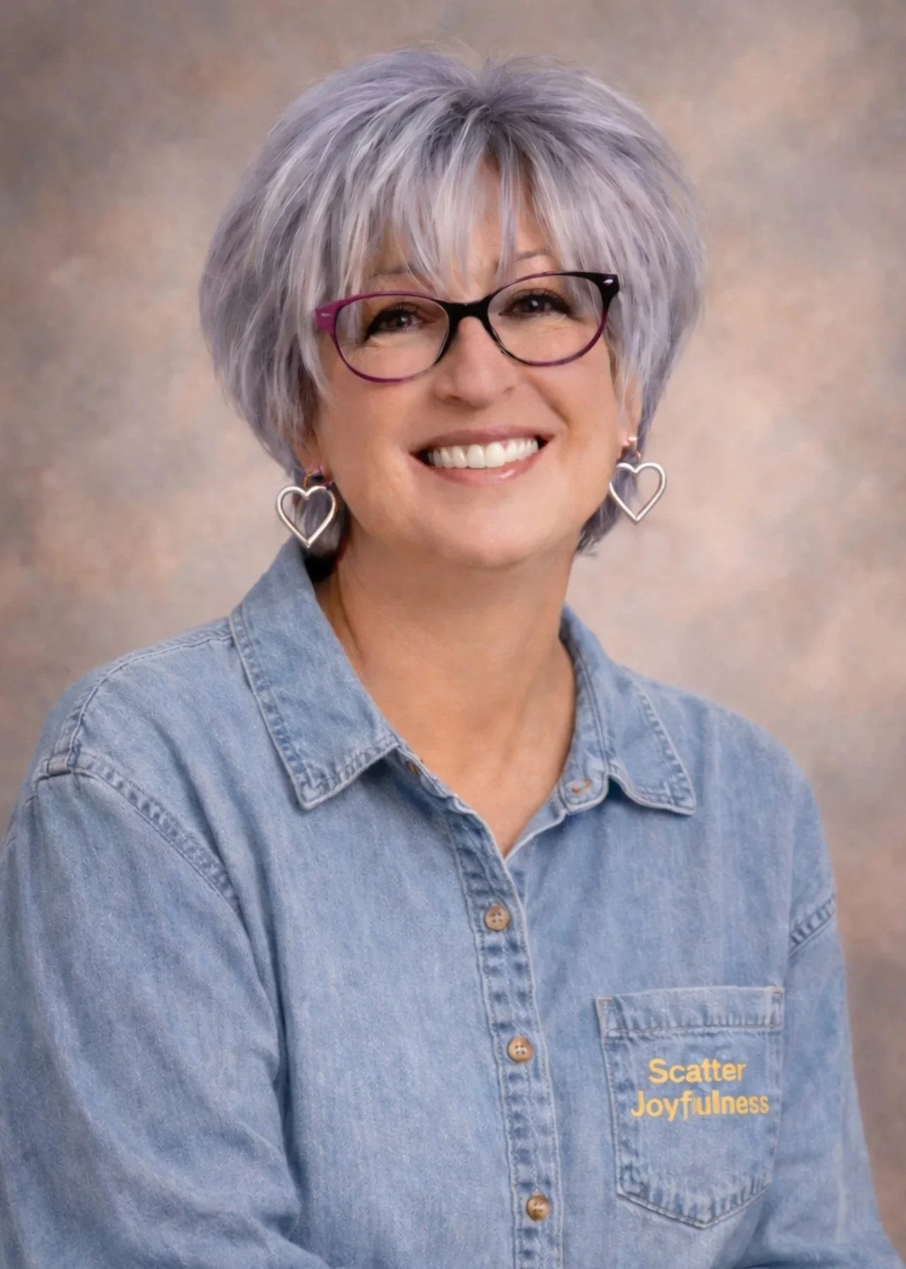 Founder portrait of Joy smiling with short silver hair and glasses, wearing a denim shirt with Scatter Joyfulness detail, warm and welcoming brand image