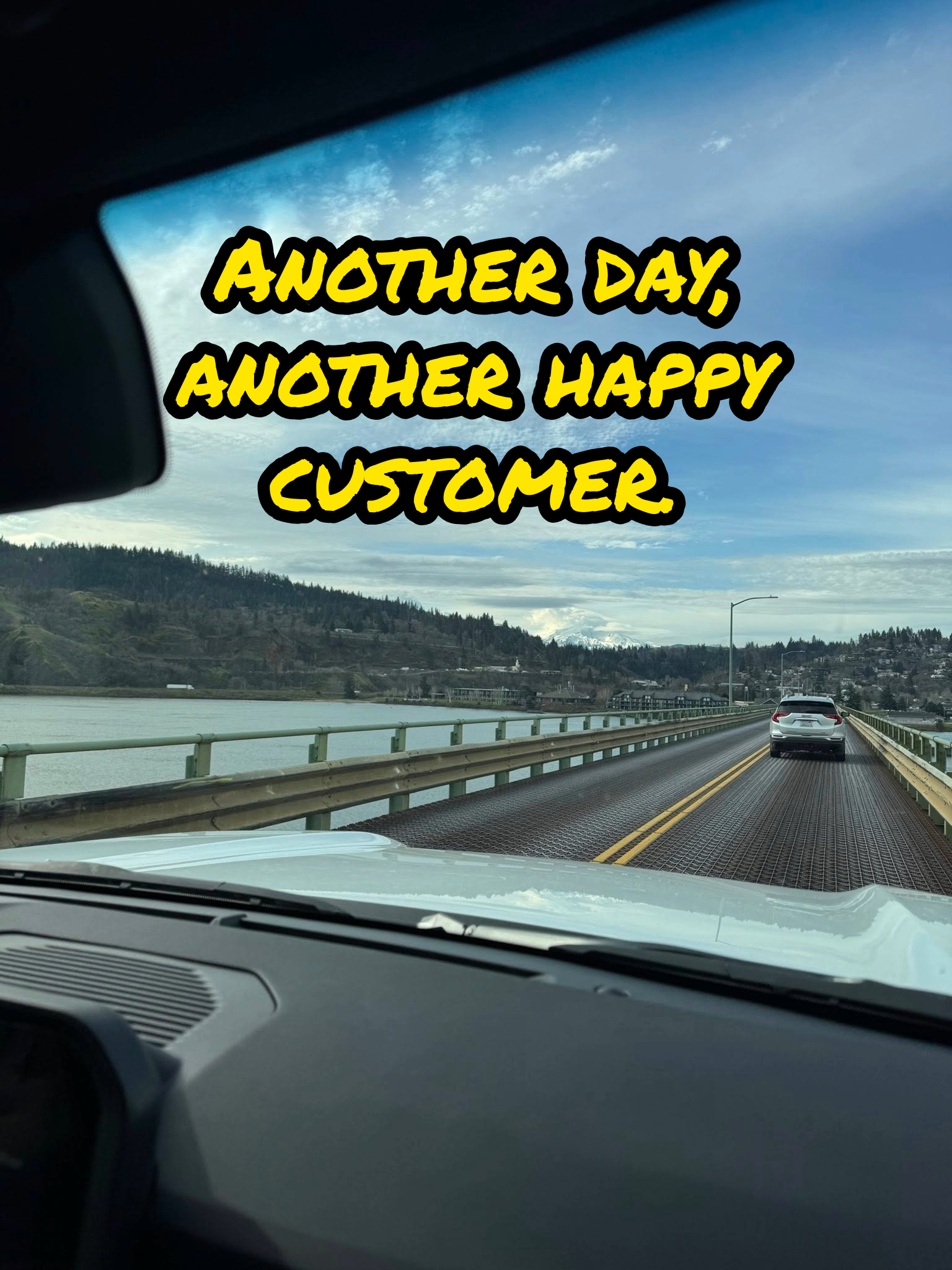 View from inside a vehicle looking out onto a bridge over water, with a car ahead and a scenic landscape with hills and clouds in the sky.