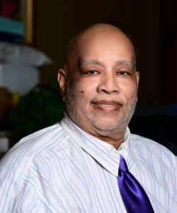 A middle-aged African American man with a bald head, wearing a white striped dress shirt and a purple tie, smiling in an indoor setting.