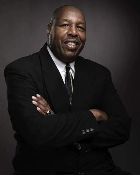 A smiling African American man in a black suit with a white shirt and black tie, posing with arms crossed against a dark background.