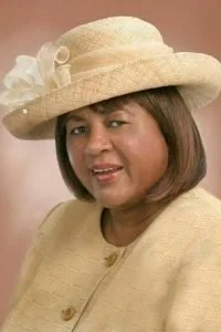 Woman wearing a light-colored wide-brimmed hat with decorative flowers, dressed in a matching light-colored outfit, smiling softly.