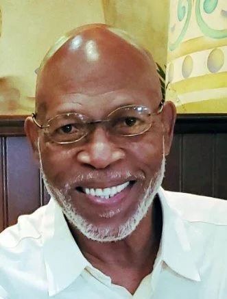 Close-up of smiling older man with glasses and a beard, wearing a white shirt in a restaurant or cafe setting.