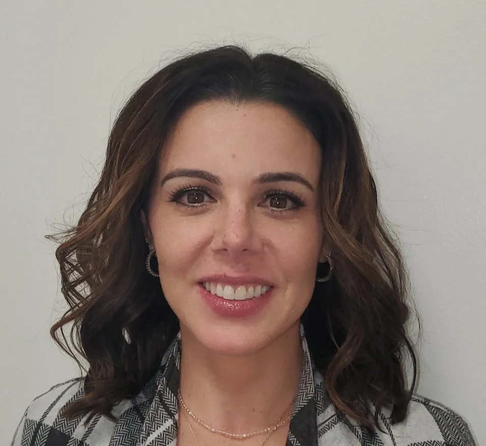 A woman with shoulder-length wavy brunette hair, wearing makeup, earrings, a necklace, and a patterned blazer, smiling at the camera against a plain white wall background.
