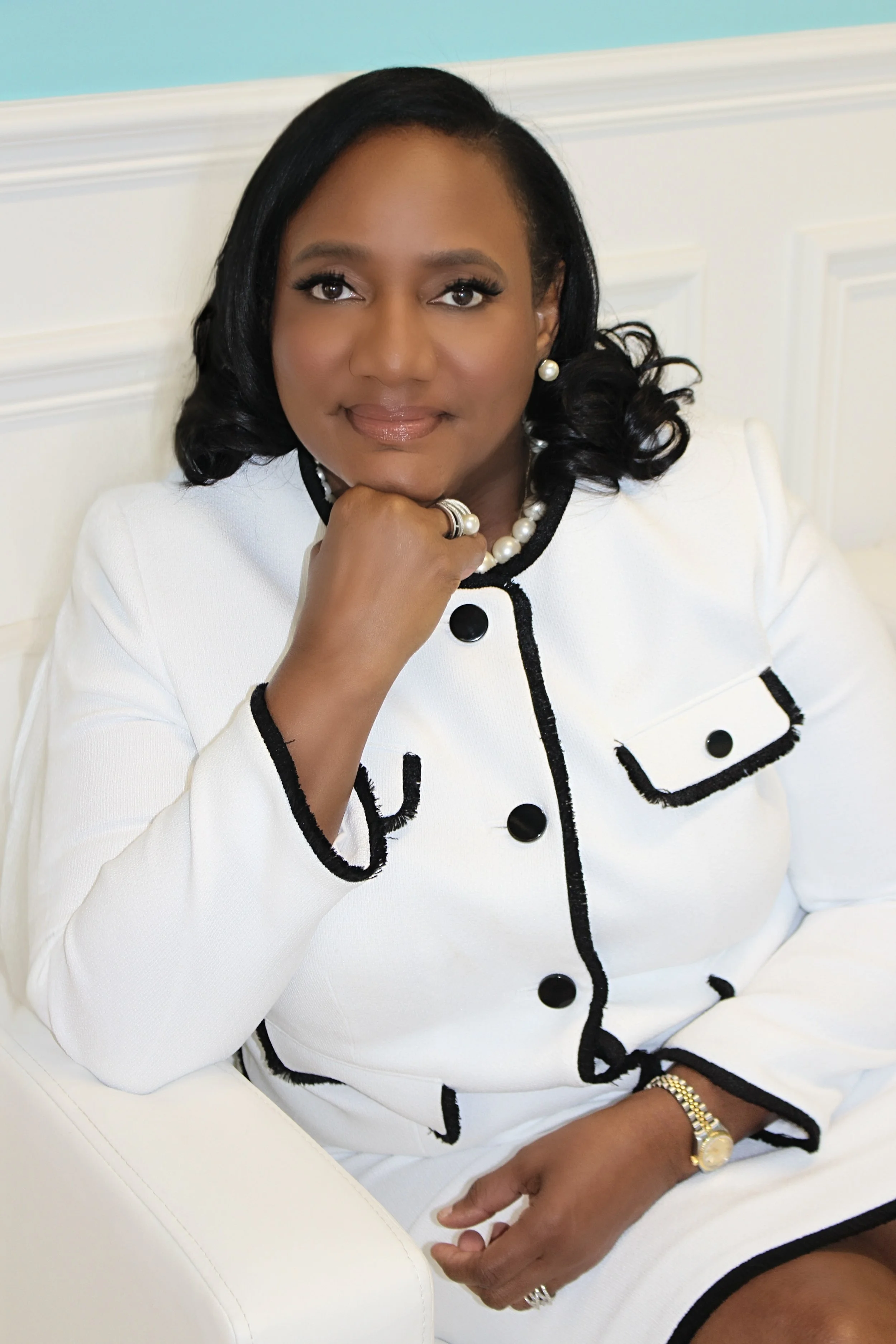 A woman with dark hair sitting on a white chair, wearing a white jacket with black trim, pearl jewelry, and a gold watch, resting her chin on her hand.