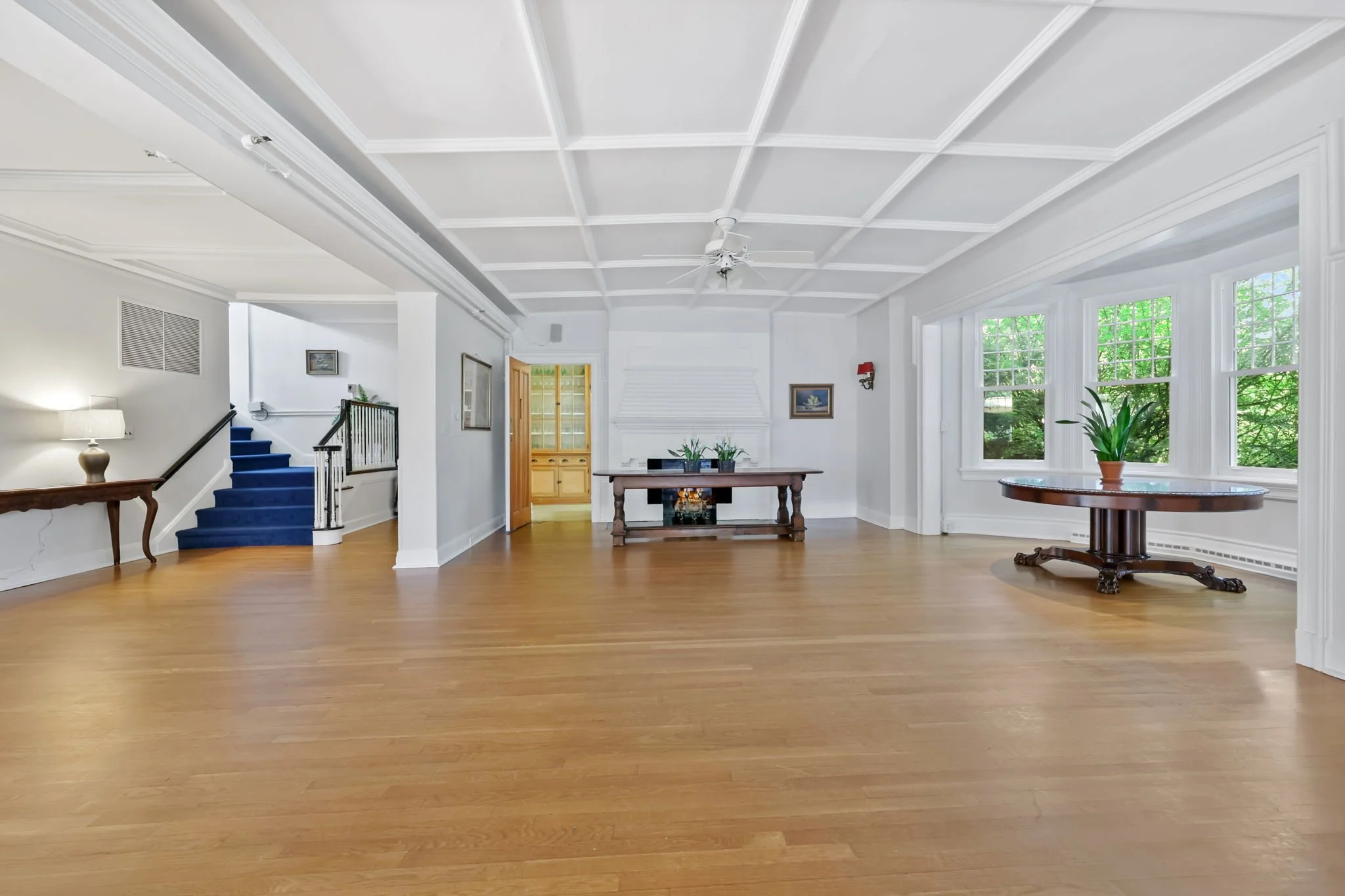 Spacious living room with hardwood floors, white walls, large windows with greenery outside, a wooden table with a potted plant, a large table at the back, and a staircase with blue carpeted steps.