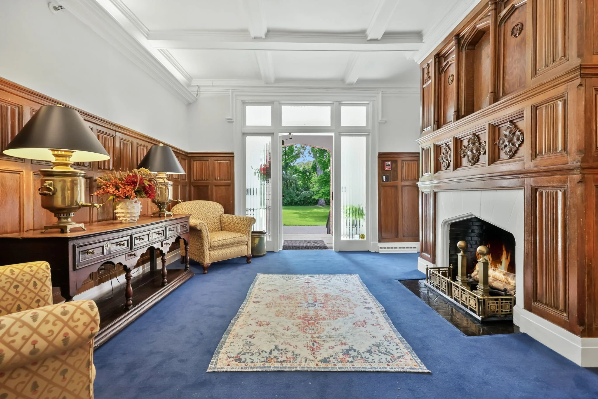 A cozy living room with blue carpet, wooden wall paneling, a white ceiling with beams, a fireplace with a fire, a rug, and a view of a garden through glass doors.