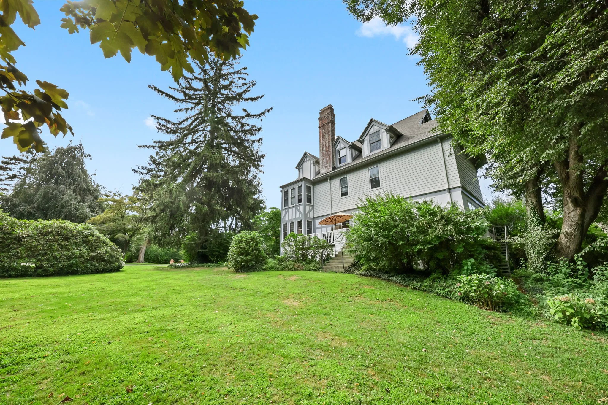A large white house with multiple gabled roofs and a tall brick chimney, surrounded by lush green trees and a well-maintained lawn on a sunny day.