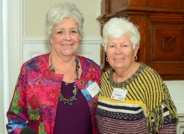 Two elderly women smiling and standing together indoors. One wears a colorful patterned blazer and necklace, the other a striped sweater with fringes. They both have short white hair and are wearing name tags.