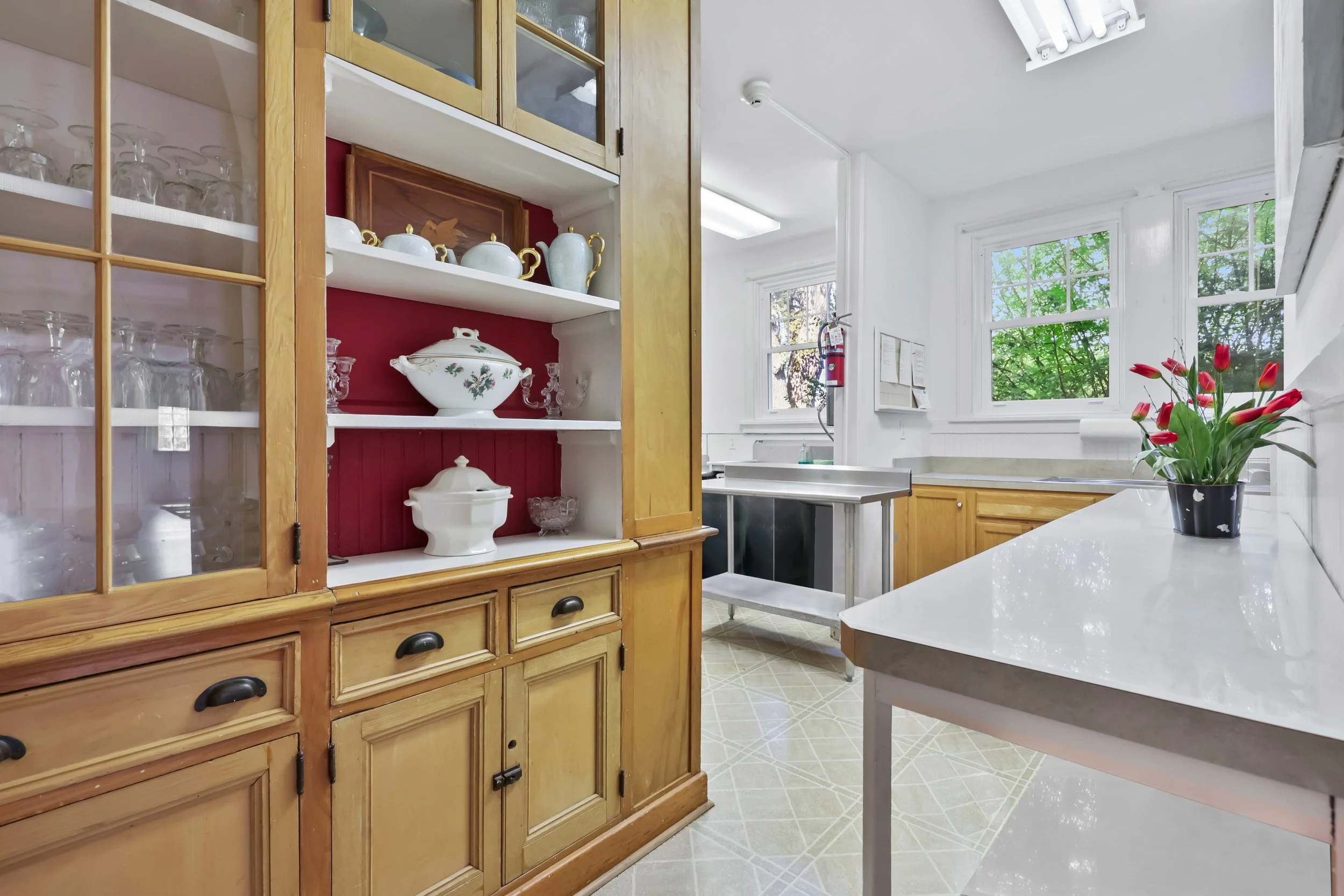 A kitchen with wooden cabinets, glassware, and a flower pot with red tulips on a white countertop. Large windows let in natural light, showing green trees outside.