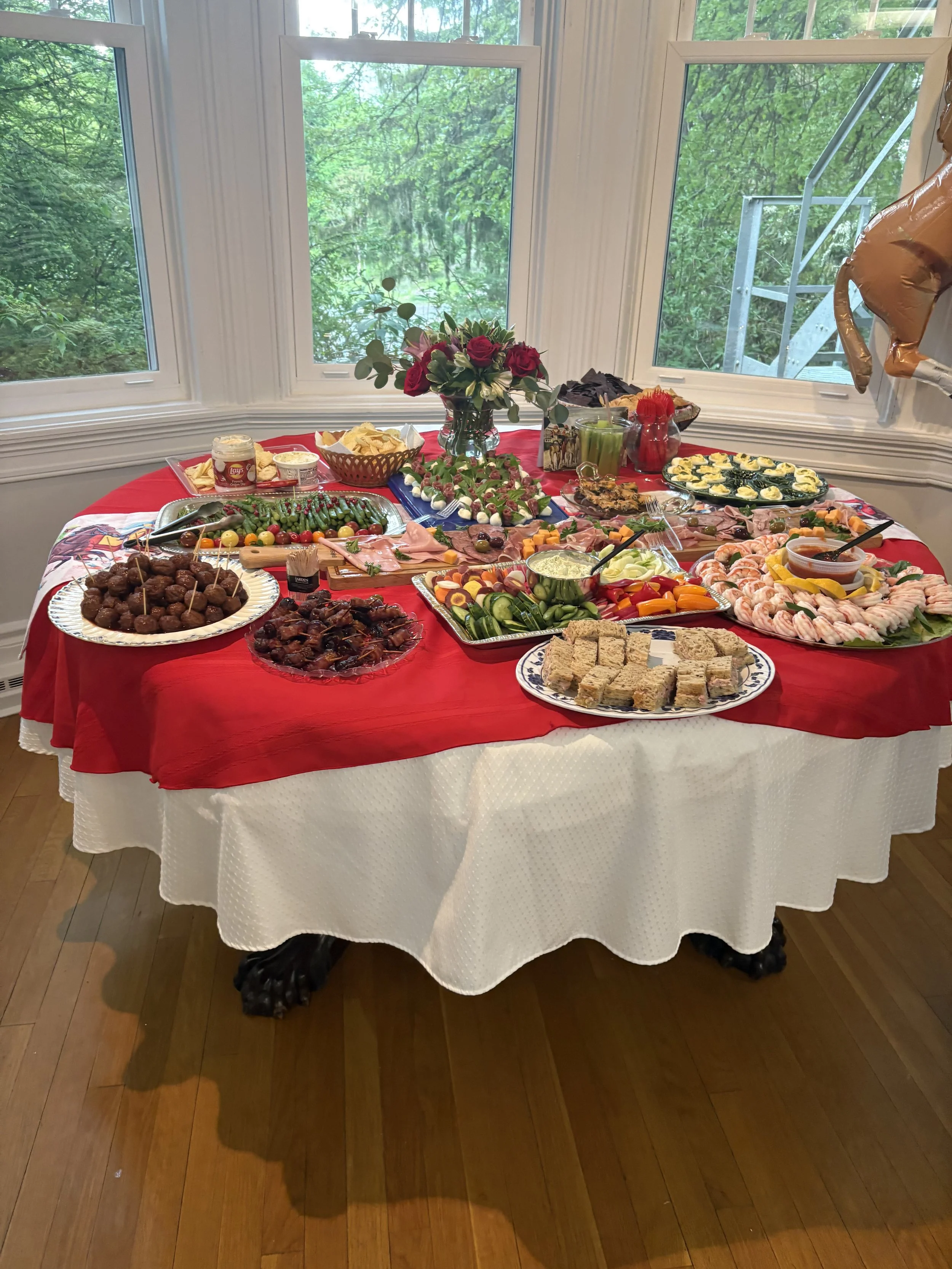 A table set with various appetizers and snacks, including meatballs, vegetable platter, shrimp platter, finger sandwiches, and dips, decorated with a floral centerpiece, with trees visible outside through large windows.
