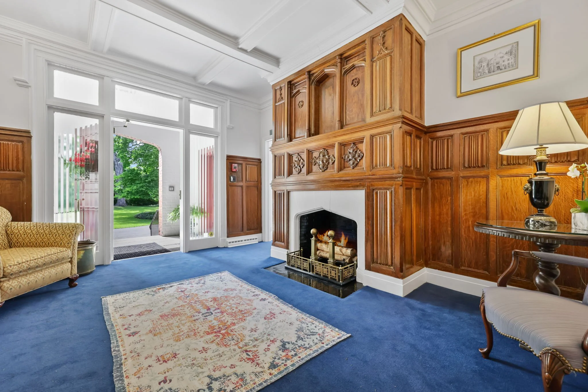 Living room with blue carpet, wooden-paneled walls, large fireplace, gold and white rug, armchair, side table with a lamp, open door leading to outdoor garden.
