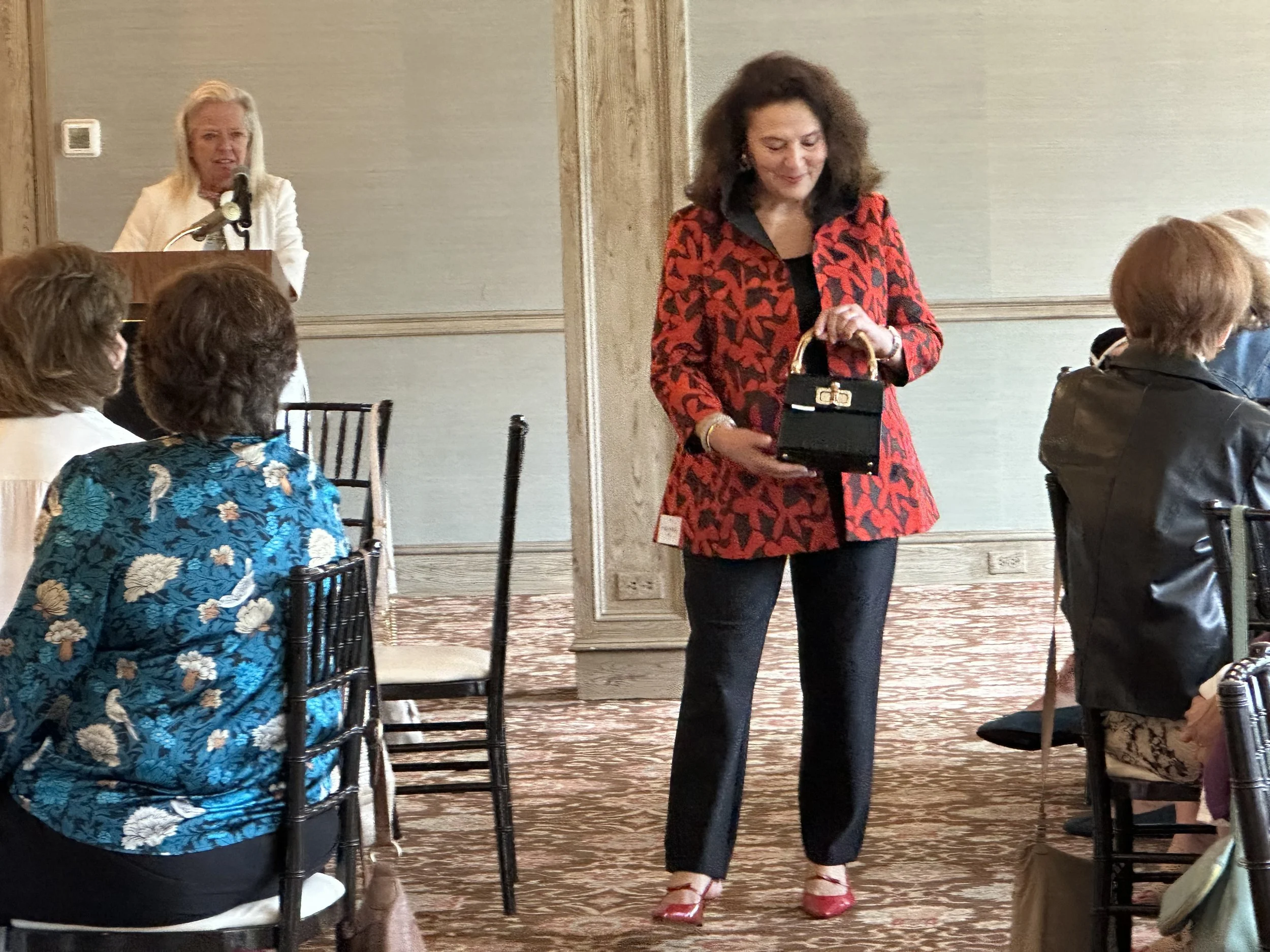 A woman in a red and black patterned blazer holding a small black handbag stands in a conference room with seated women and a woman at a podium in the background.
