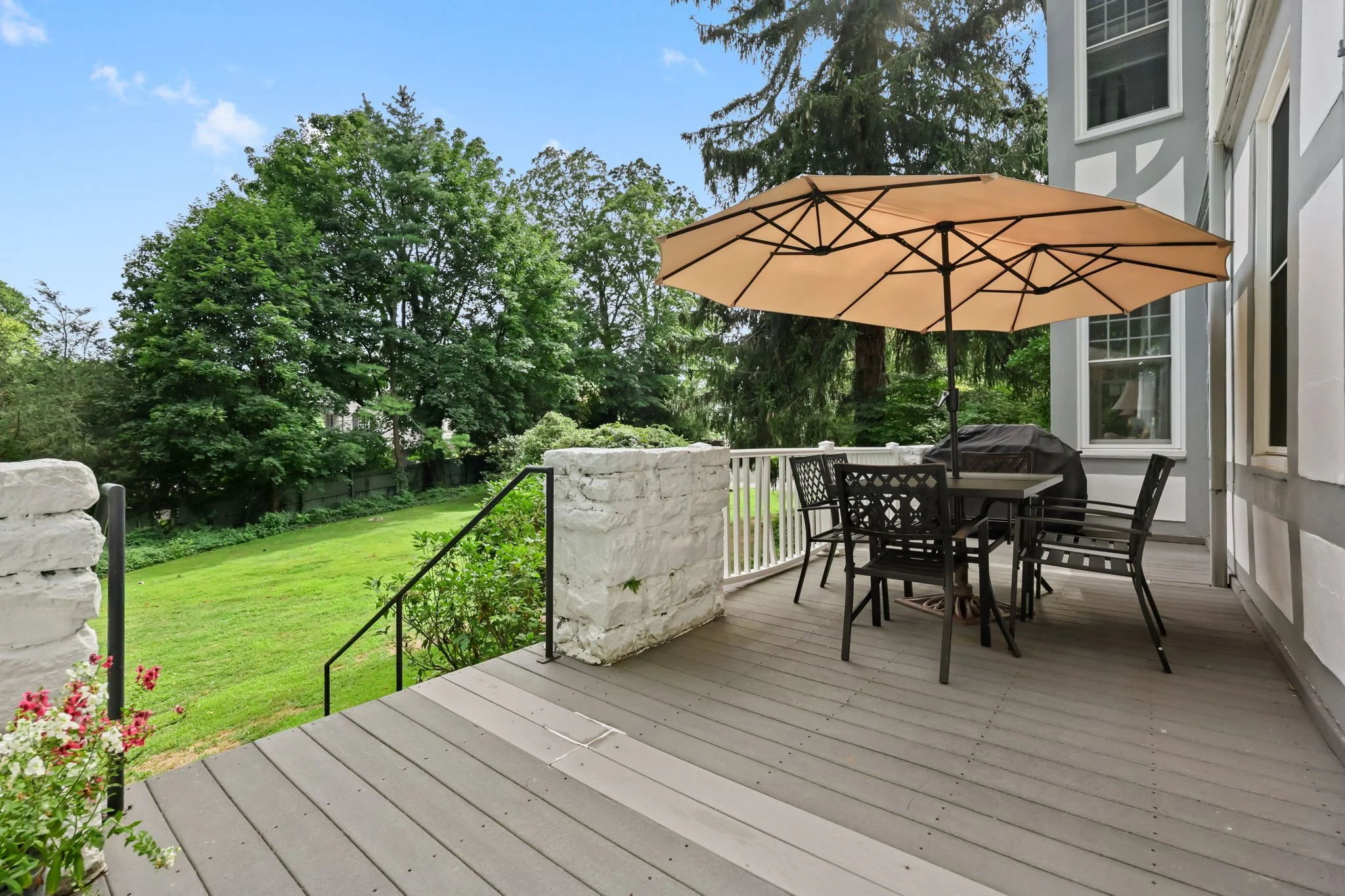 Back porch with outdoor dining table, chairs, large umbrella, and a grill, overlooking a grassy backyard with green trees.
