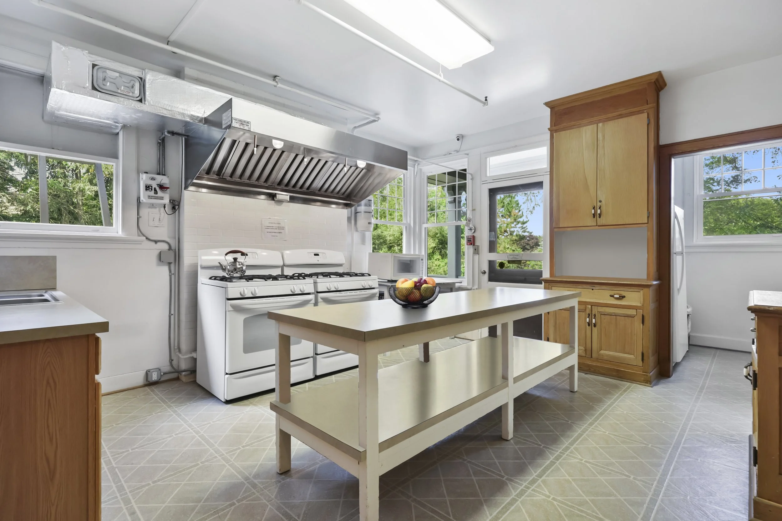 Kitchen with white stove, vent hood, wooden cabinets, a white island with fruit basket, and multiple windows with greenery outside.