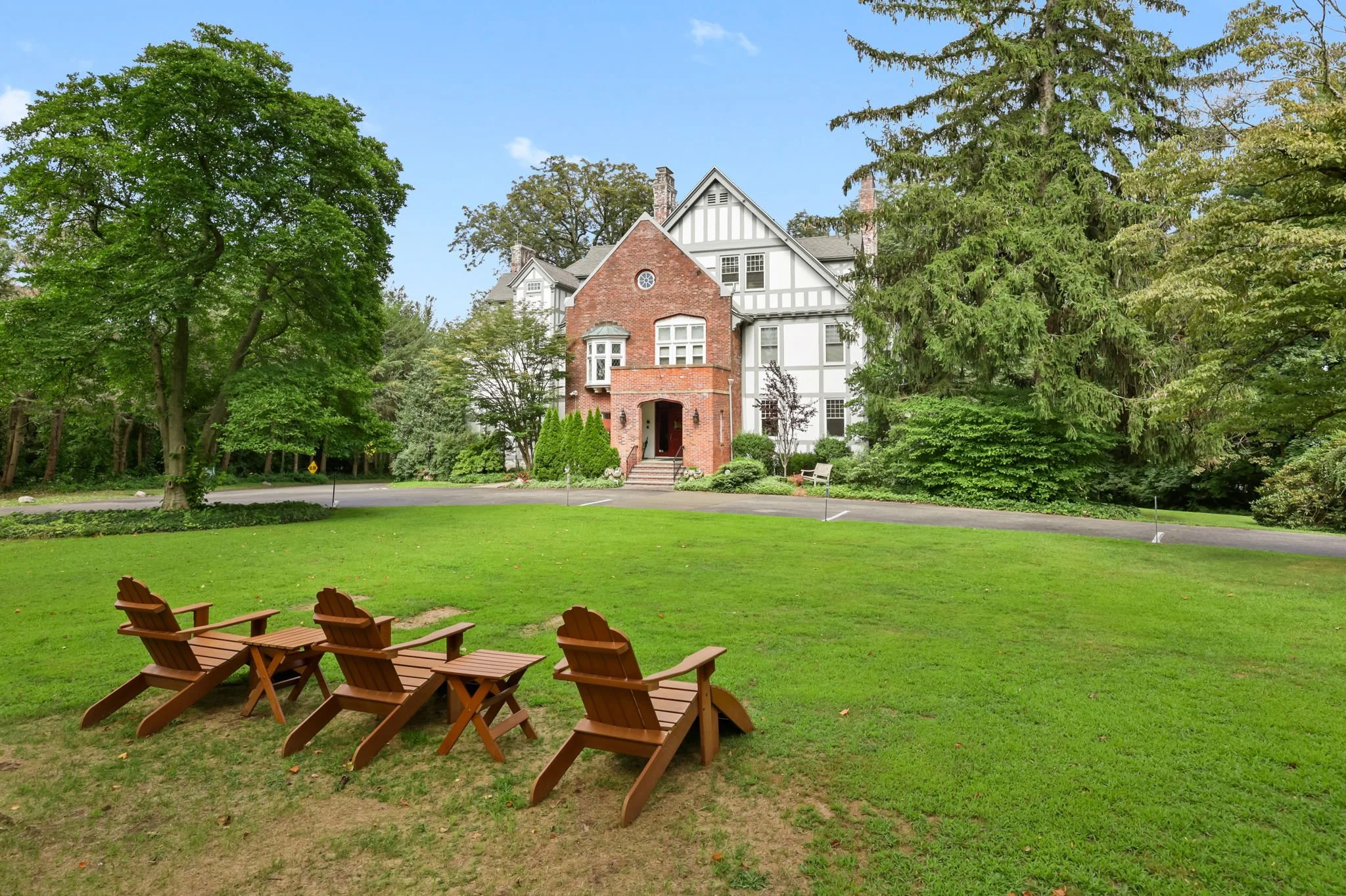 A classic house with a brick front, white walls with black trim, surrounded by trees and greenery. In the foreground, four wooden Adirondack chairs are arranged on a grassy lawn.