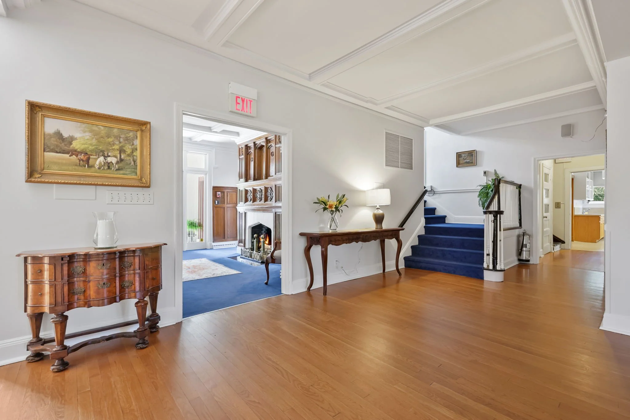Interior of a house with a wooden floor, a staircase with blue carpet, a side table with a vase and lamp, a hallway leading to a kitchen, and a room with a fireplace and blue carpet.