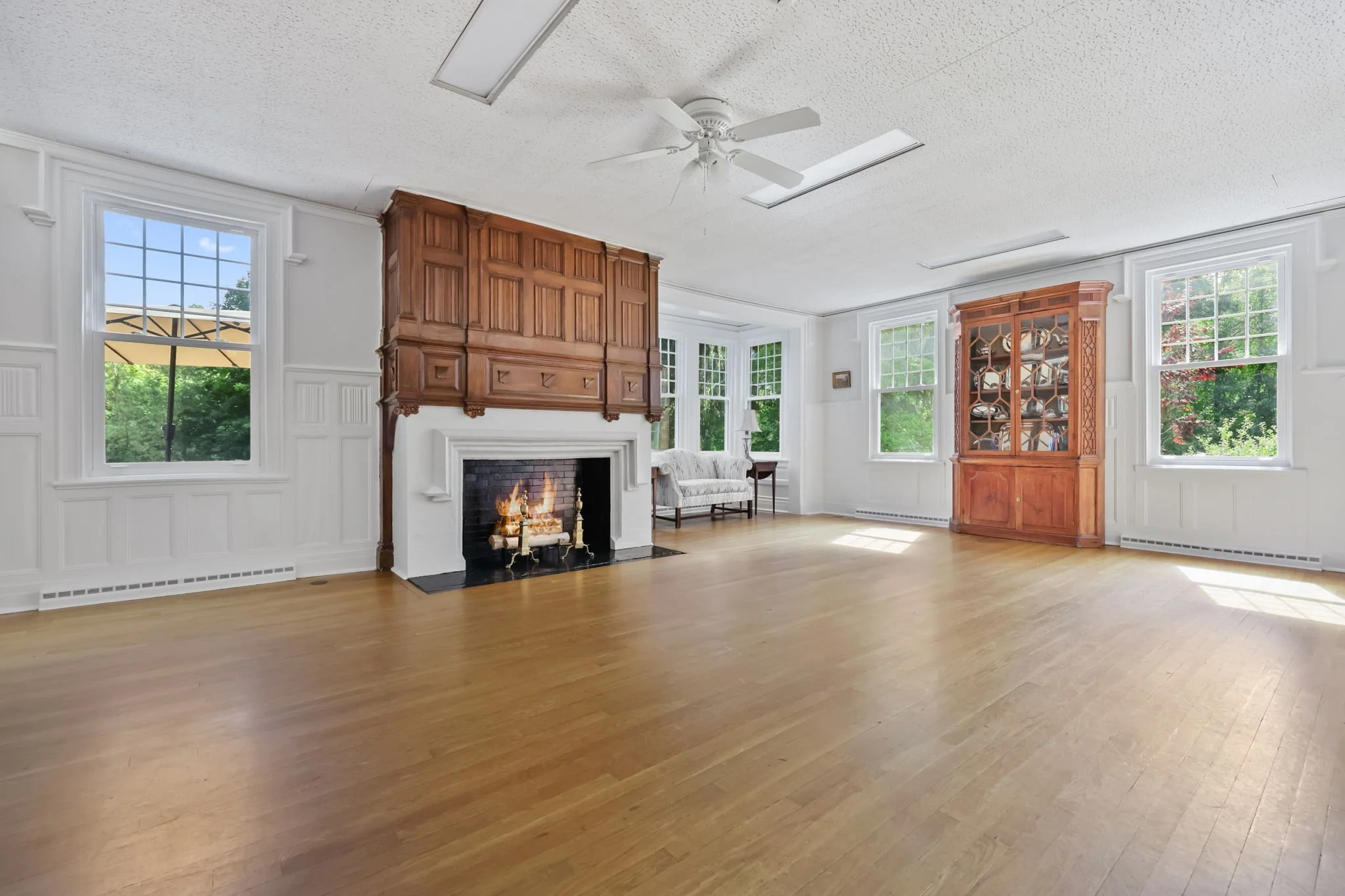 Spacious living room with hardwood floors, white walls, multiple windows, a fireplace with a wood mantel, a white sofa, and a wooden cabinet with glass doors.