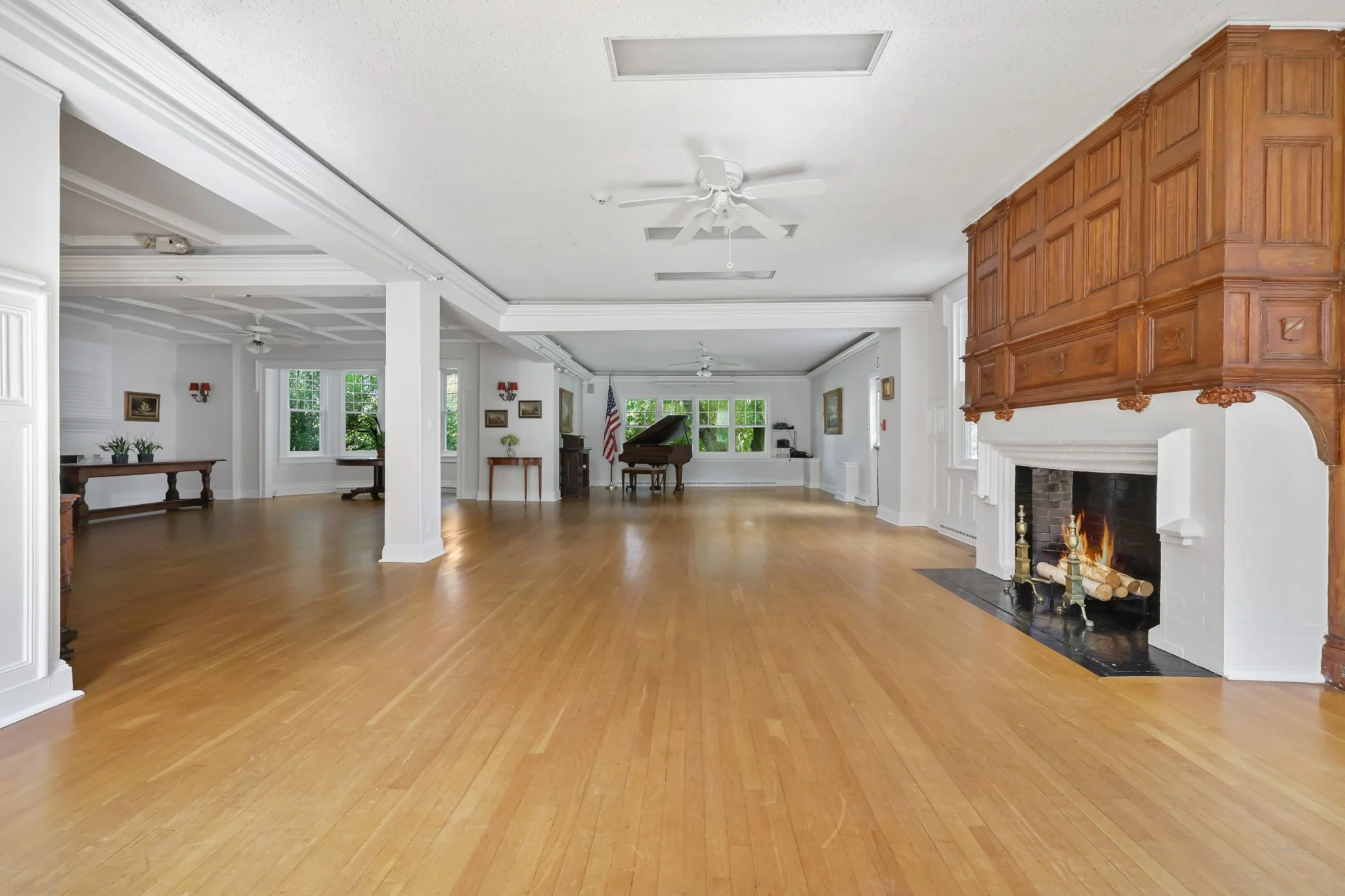 Spacious living room with white walls, wooden flooring, a brick fireplace with wood logs, ceiling fans, and large windows showing trees outside.