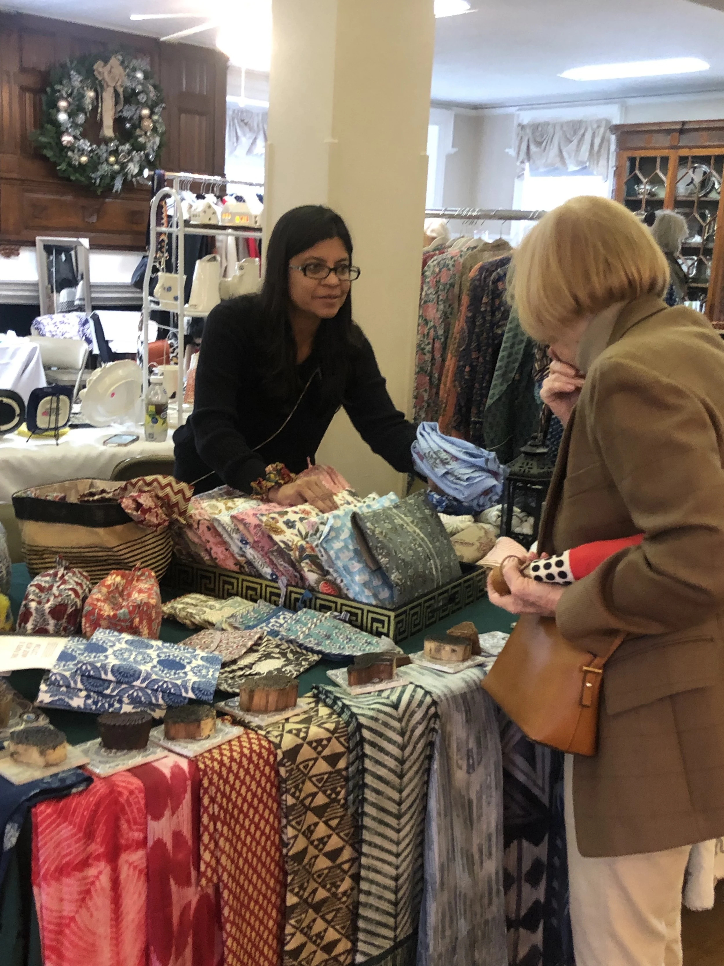 A woman with glasses and dark hair is selling colorful fabric and small handmade items at a craft fair. She is interacting with a customer who is wearing a tan blazer and holding a polka dot item.
