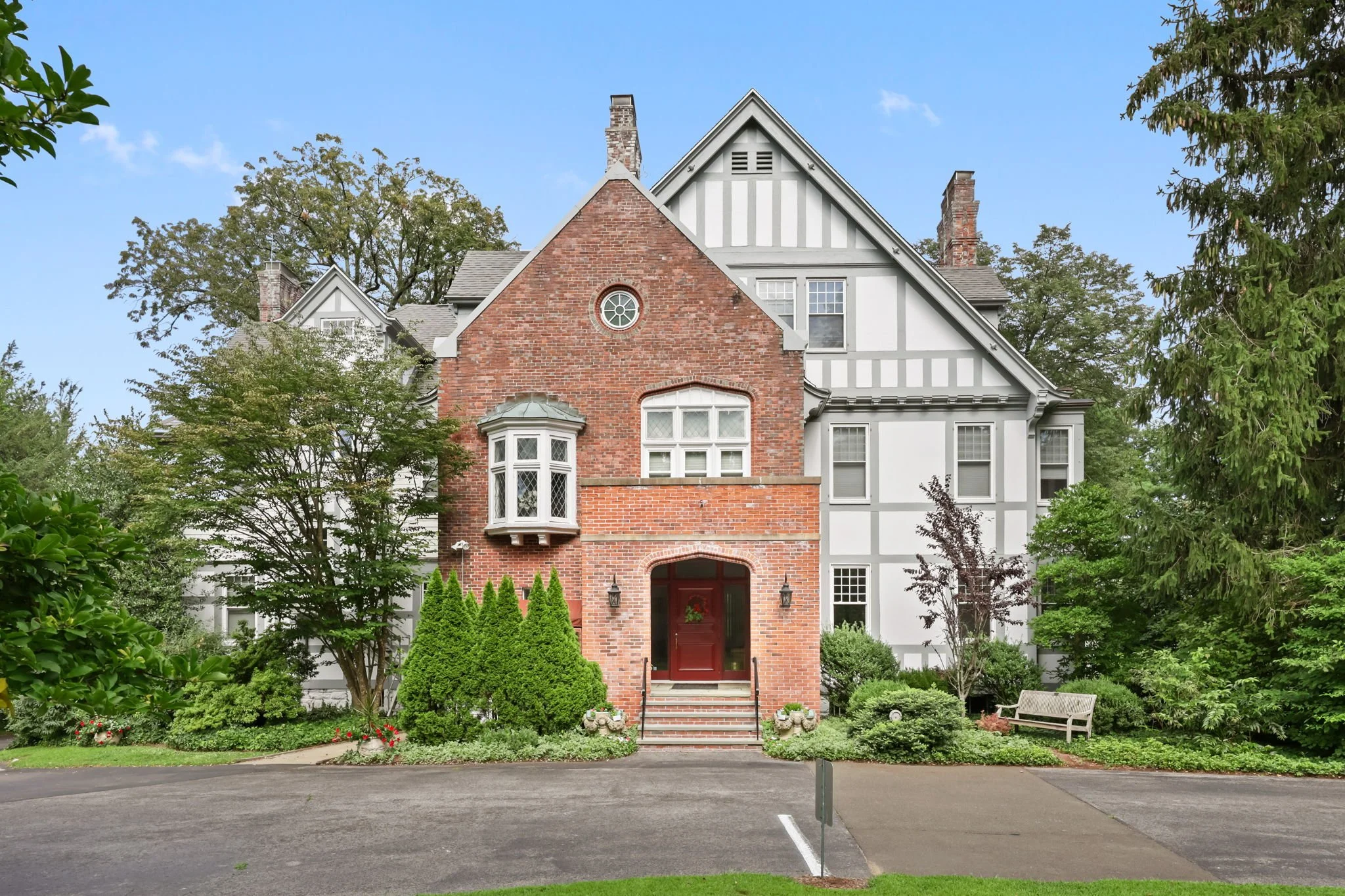 Front view of a large, historic house with a brick and white facade, multiple windows, and a red front door, surrounded by lush greenery and a paved driveway.