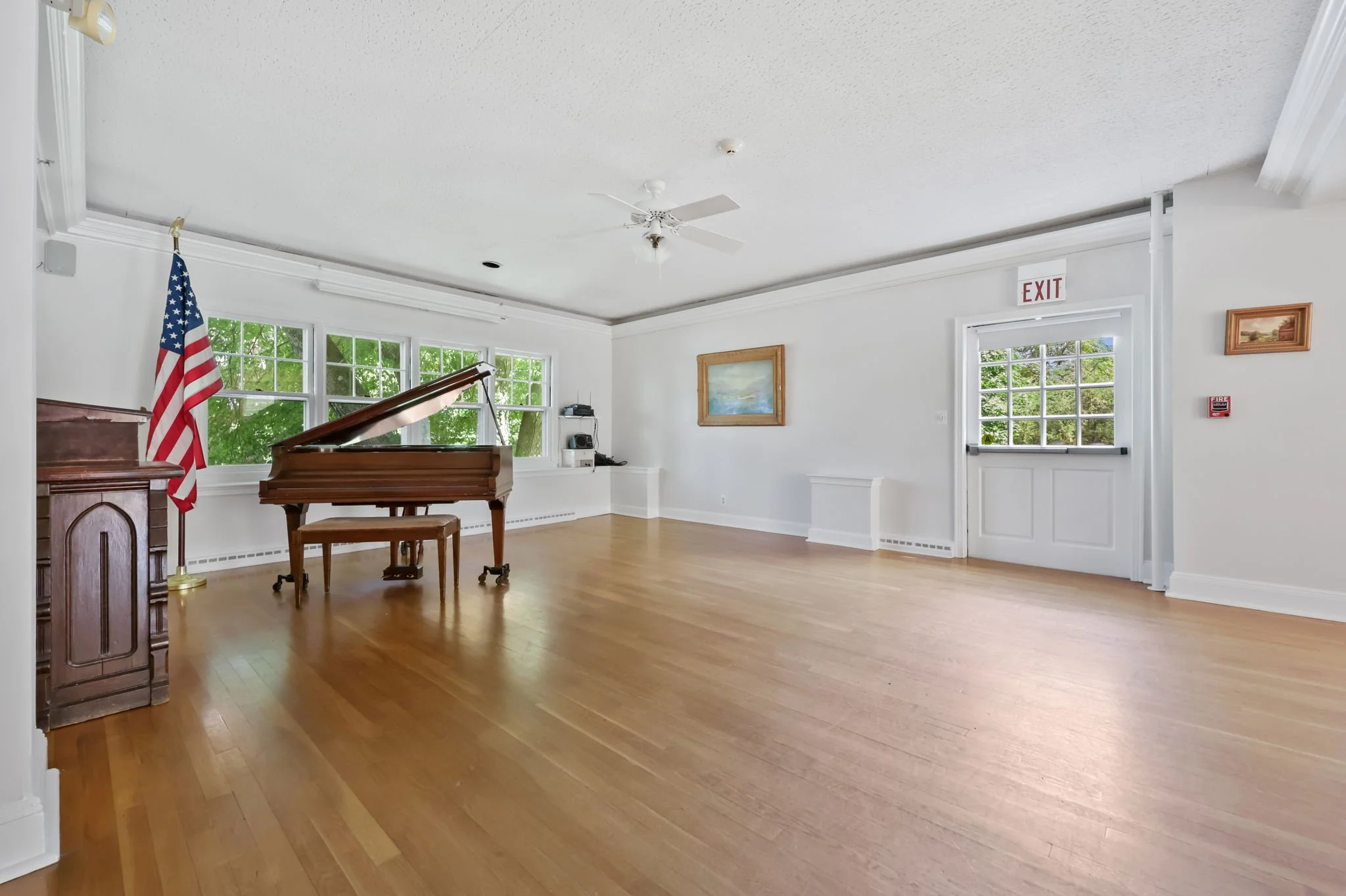 Empty room with wooden floors, white walls, large windows, a grand piano, an American flag, a ceiling fan, and wall decorations.