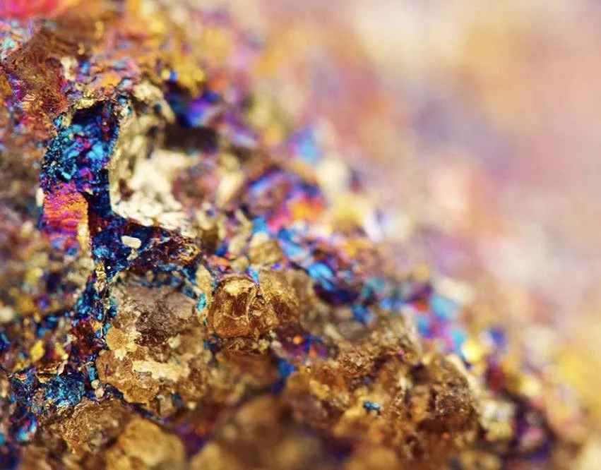 Close-up of colorful mineral or metallic substance, showing vibrant blue, pink, purple, gold, and brown hues with textured, rough surface.