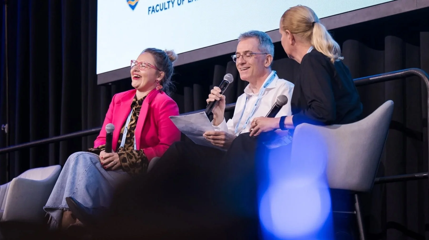 Three people sitting on stage at a conference, engaged in discussion. The woman on the left is wearing glasses, a bright pink blazer, and a leopard print top, and is holding a microphone. The man in the middle is smiling, holding a piece of paper, and speaking into a microphone. The woman on the right is turned towards him, with blonde hair tied back, and is listening attentively. The background features a large screen displaying a logo and text.