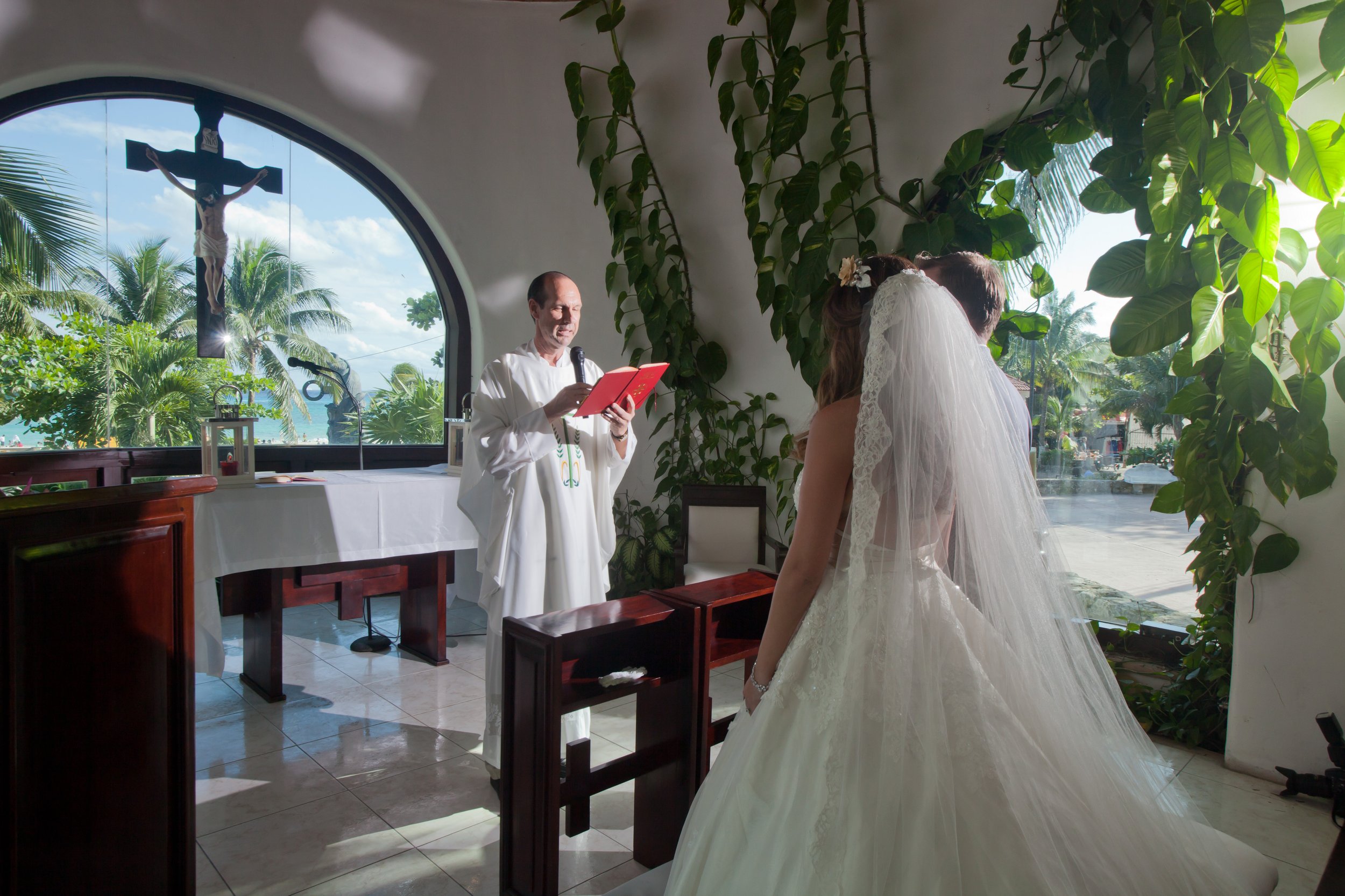 A bride dressed in a white wedding gown and veil standing in front of a priest during a wedding ceremony inside a chapel with large arched window, greenery, and sunlight.