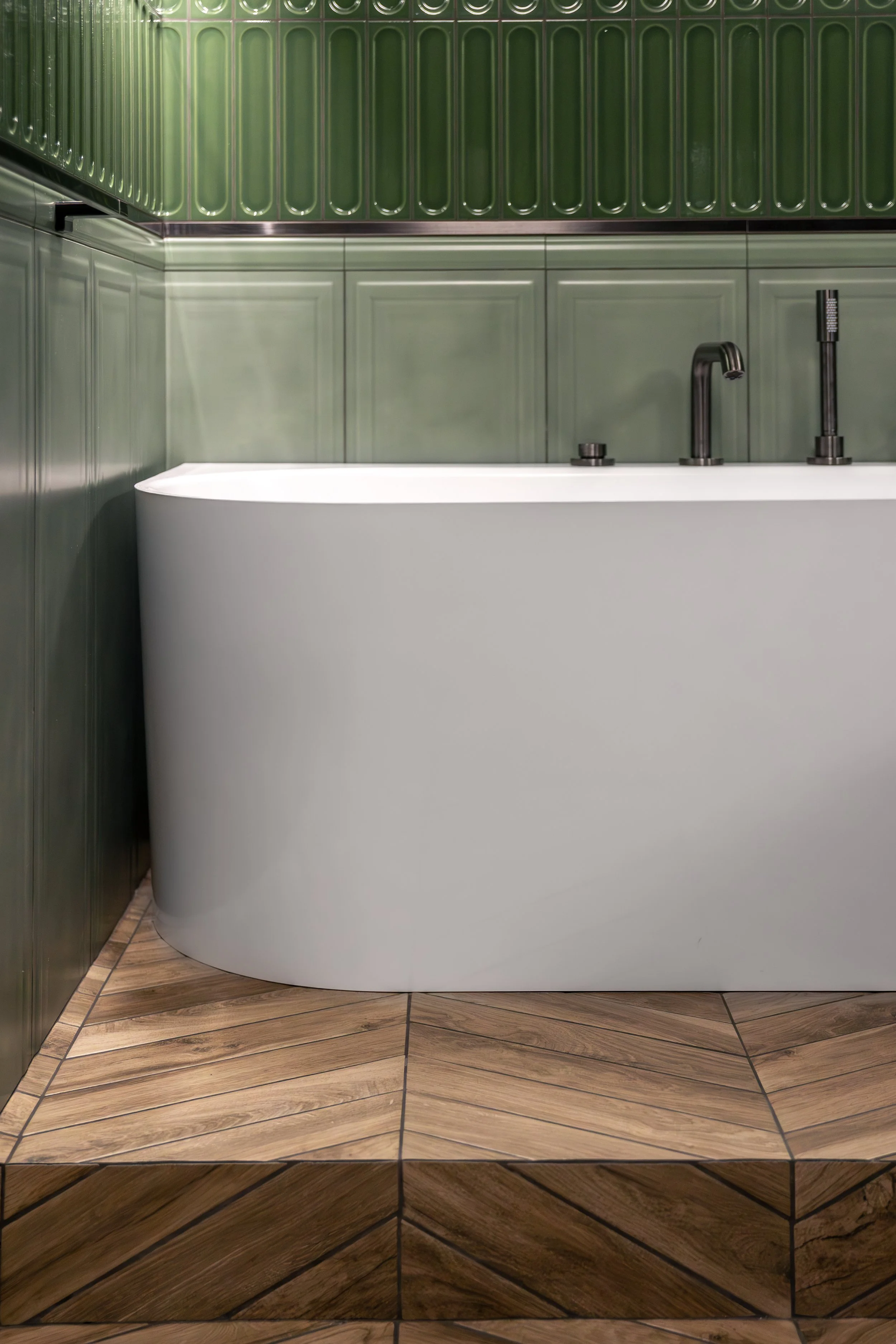 Modern bathroom design with green tiled walls, a white curved bathtub, and wooden floor tiles.