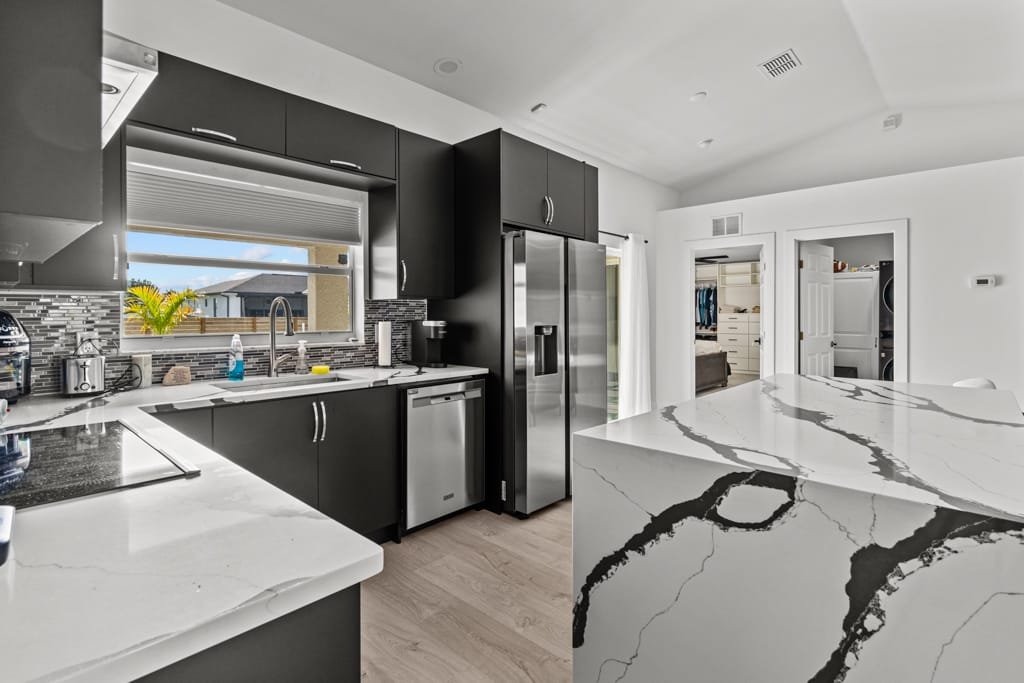 Open concept, modern kitchen design with black cabinets, stainless steel appliances, marble island, gray backsplash, and a window with a view.